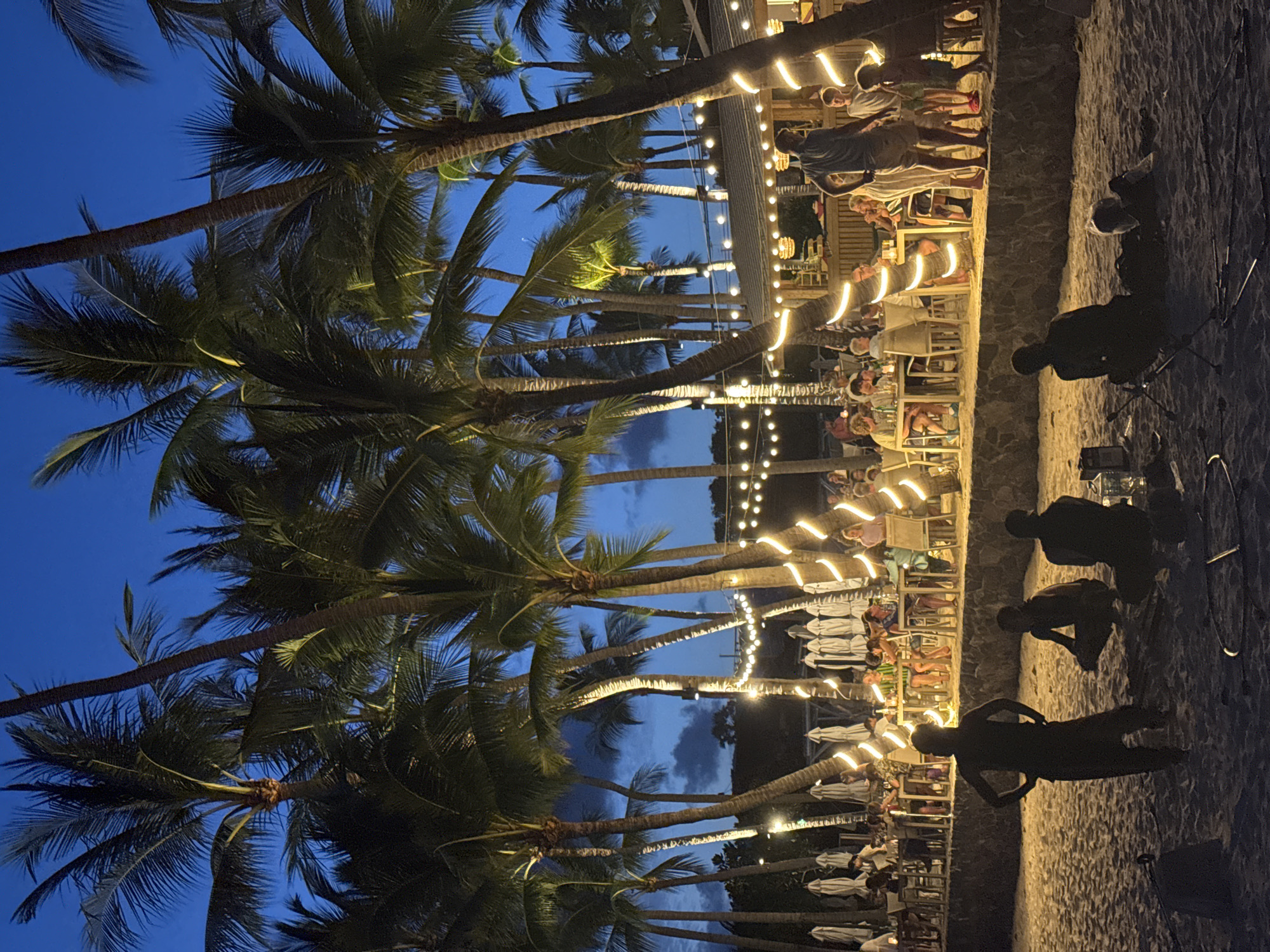 The image depicts a tropical beachside restaurant scene at dusk in Ko Samui District, Thailand. The foreground shows a sandy beach with three silhouetted figures, one standing with hands on hips and two others seated or crouching. In the midground, a raised stone wall separates the beach from a dining area where numerous people are seated at tables under palm trees adorned with string lights. White umbrellas are positioned behind the tables. In the background, more palm trees are visible against a dark blue twilight sky. The atmosphere appears lively and festive, with the lighting suggesting it is evening.