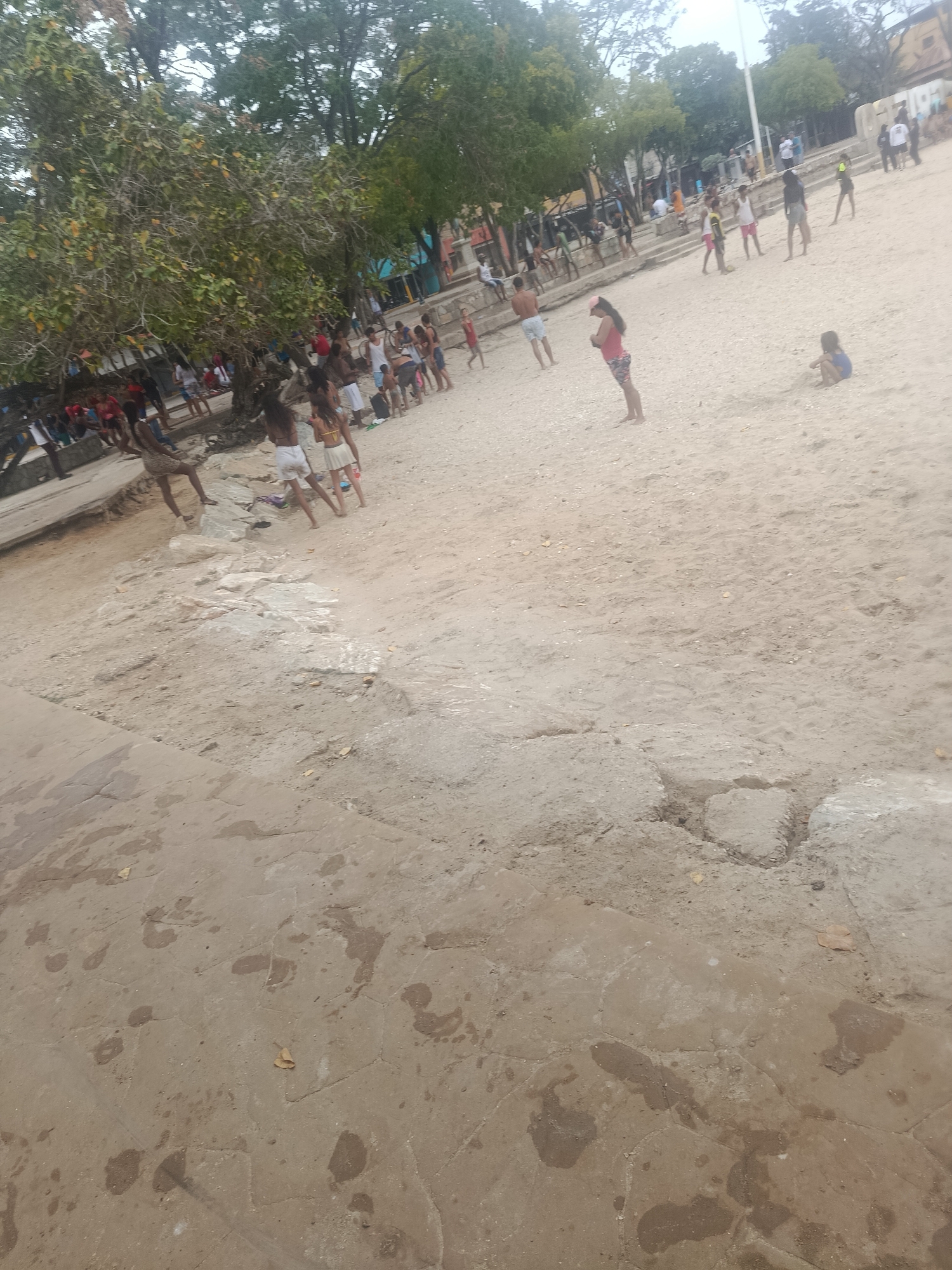 In Juan Griego, Venezuela, a beach scene unfolds with numerous people scattered across a sandy expanse under a canopy of trees. The foreground displays a concrete area with numerous footprints and water stains. Further back, a crowd of individuals, varying in age and attire, engage in leisure activities. Several people are seen walking on the sand, while others are seated. Some individuals appear to be playing a game on the beach. In the mid-ground, a woman in a pink top and patterned shorts stands with her back to the camera, facing away from a group of people. In the background, more people are visible, as are structures that suggest the presence of buildings and possibly a promenade. The overall environment suggests a lively public space, likely a beach or park area, where people are enjoying the outdoors.