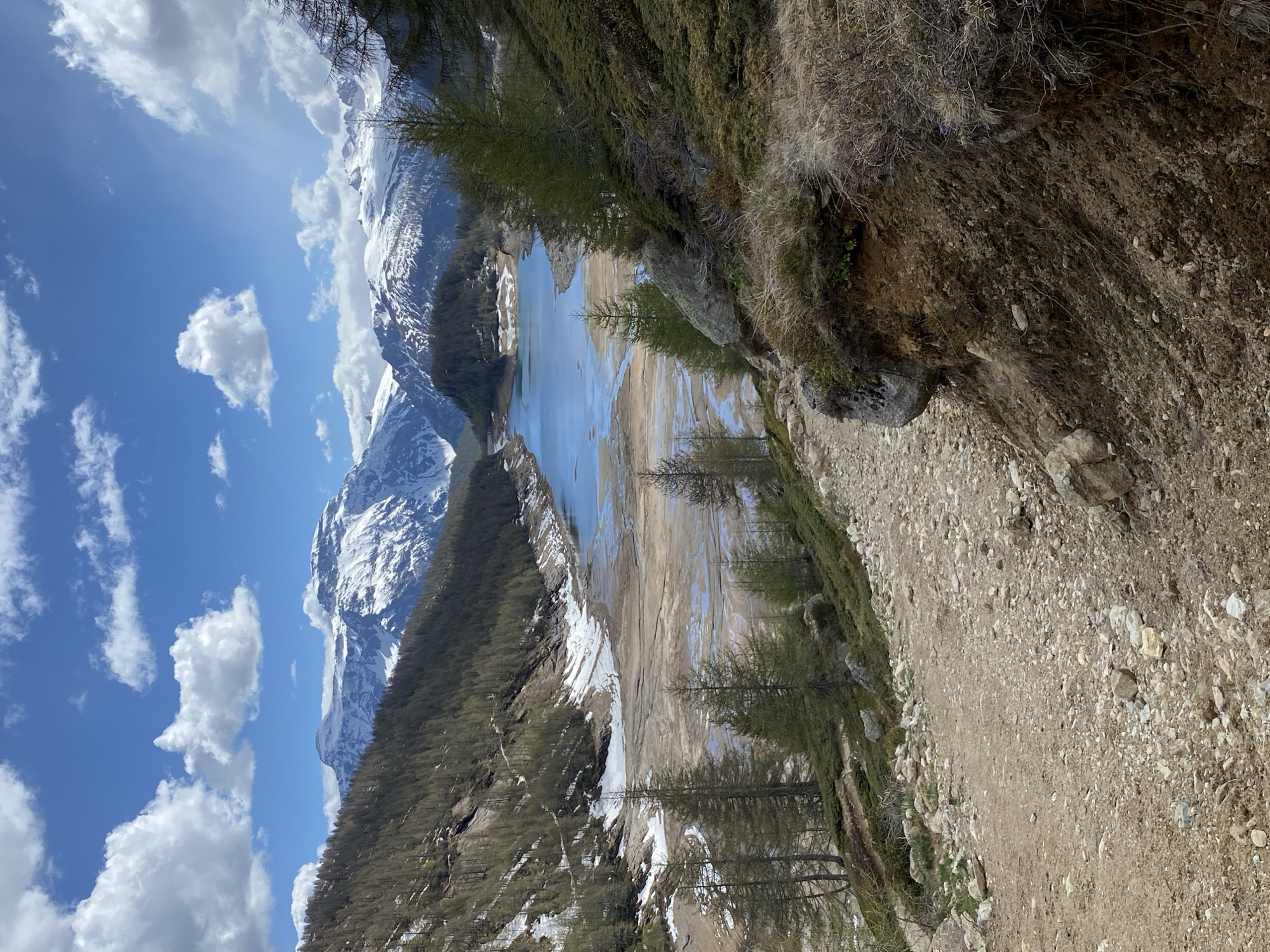 The image captures a mountainous landscape in Baceno, Italy, overlooking a lake or reservoir. The view is framed by a rocky, gravelly slope on the right, which descends toward the water. The water level appears to be low, exposing a dry lakebed or shoreline. Beyond the water, a dense forest extends up the slopes of the mountains. Snow-capped peaks rise in the background, partially obscured by clouds in a blue sky. The time of year appears to be spring or early summer, based on the combination of snow and greenery. No people are visible in the frame.