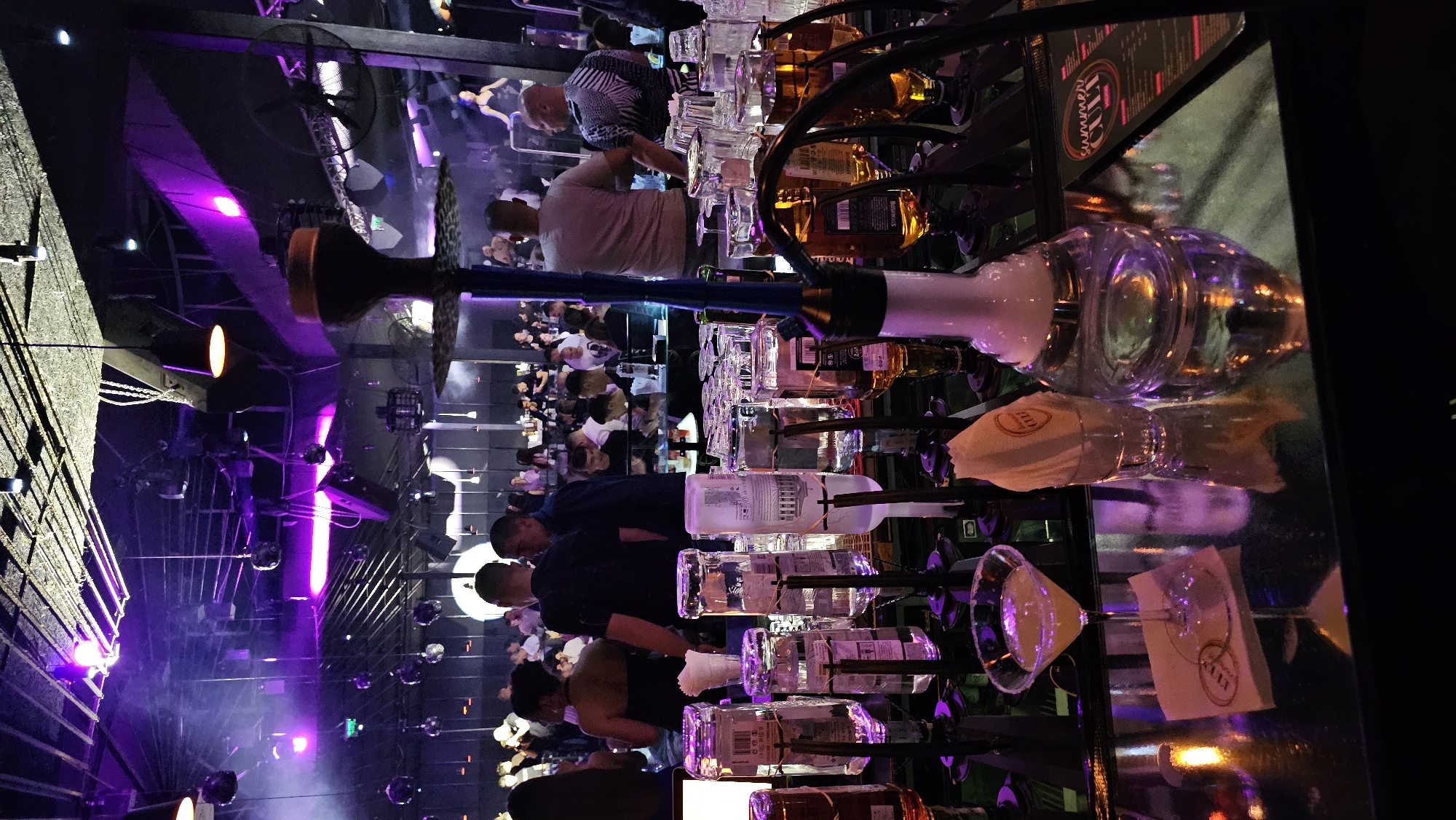 The image shows the interior of a bar or club in Varna, Bulgaria. Patrons stand at the bar and are seated at tables in the background. Bottles of alcohol are displayed on a rack behind the bar. A hookah sits on the bar counter. The bar is lit with purple lighting.