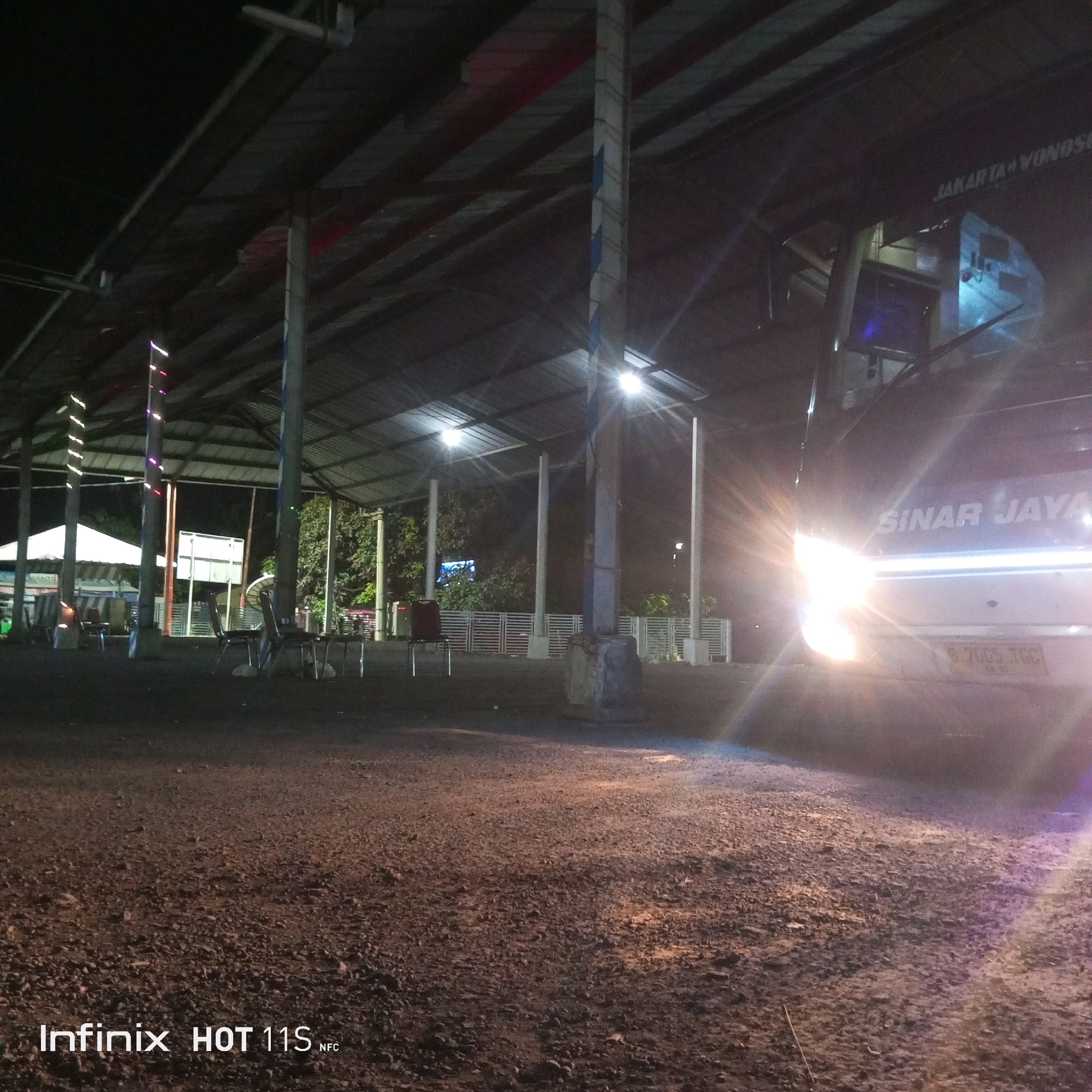 The image depicts a nighttime scene at what appears to be a bus station or rest stop in Cikawung, Indonesia. A large bus, identified by the text "SINAR JAYA" on its side, is prominently featured. The bus's headlights are on, creating a strong glare. The foreground is a gravelly, uneven ground. In the background, a covered waiting area with a corrugated metal roof is visible, supported by concrete pillars. Several chairs are arranged under this shelter, and some string lights are hanging from one of the pillars. The environment appears to be outdoors, with trees and vegetation visible beyond the covered area. The overall impression is that of a functional, albeit somewhat dimly lit, transportation hub at night.