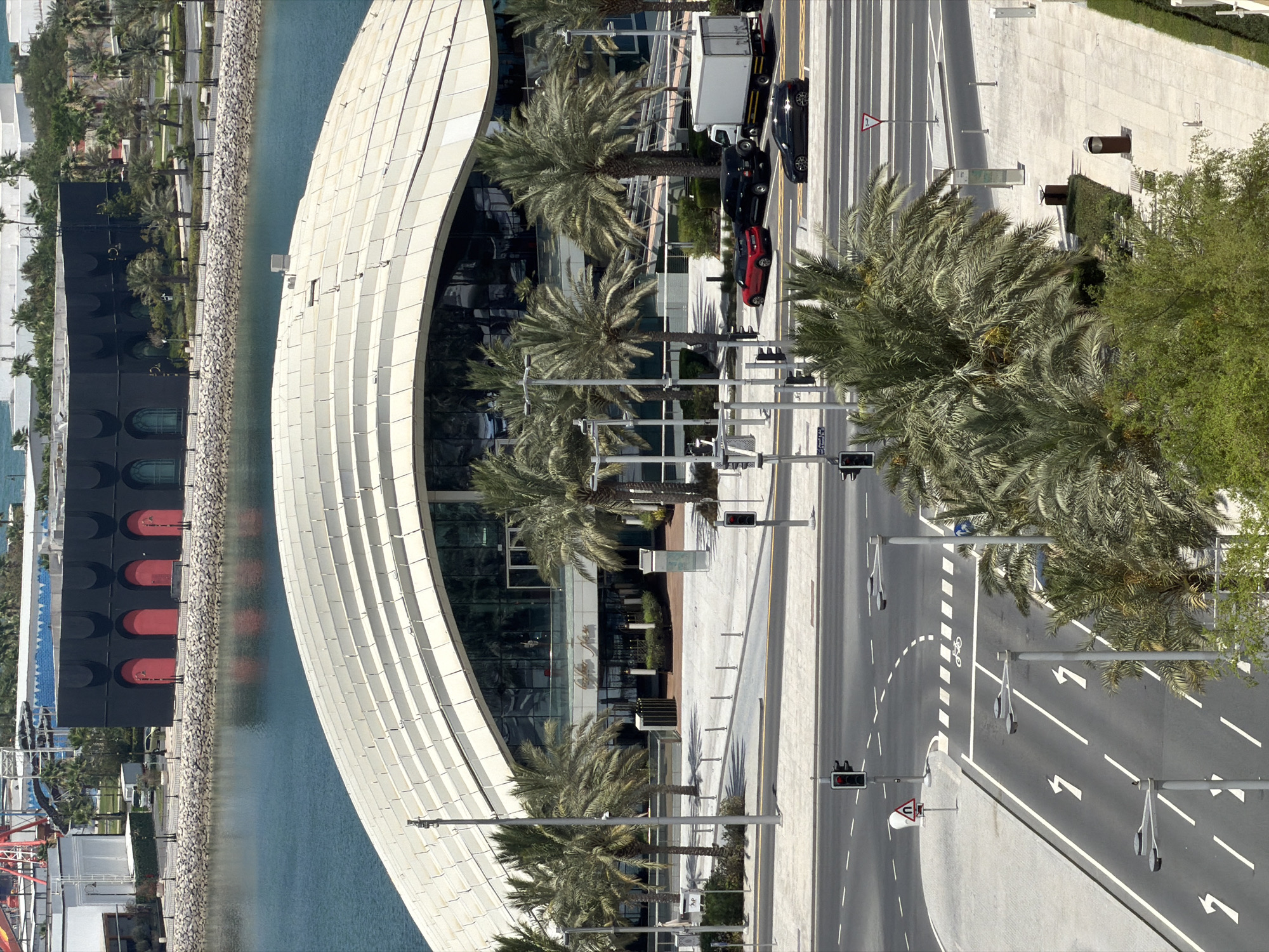 The aerial view shows a modern cityscape with a large, curved, white-roofed building in the foreground, likely a commercial or entertainment venue, with extensive glass facades. In front of this building is a street with traffic lights and several vehicles, including a red car, a black SUV, a black sedan, and a white box truck. Palm trees are scattered throughout the scene, lining the walkways and bordering the street. Beyond the main building, a body of water, possibly a harbor or canal, separates it from a district with a dark, rectangular building featuring arched red doorways. Further in the background, a construction crane is visible, suggesting ongoing development. The environment appears to be a well-maintained urban area with paved roads, sidewalks, and landscaping.