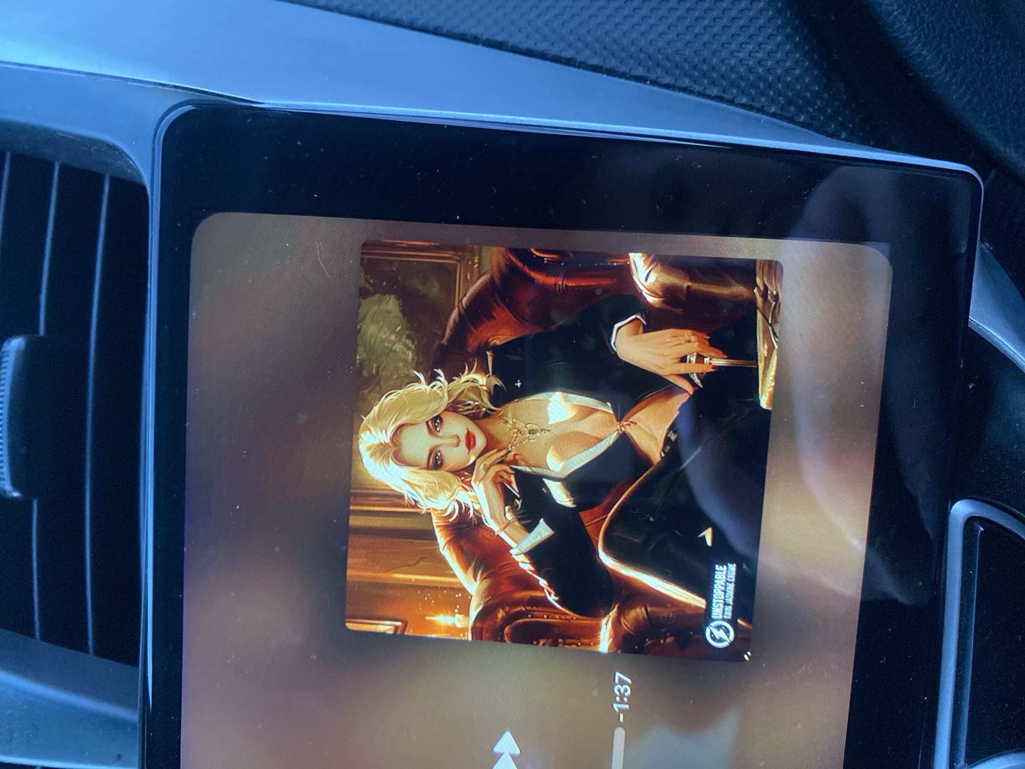 A digital screen, likely an in-car infotainment system, displays an illustrated image, possibly album art, of a seductive woman. The screen is glossy and shows reflections from the vehicle's interior and ambient light.
The central figure in the artwork is a blonde woman with fair skin and red lips, depicted in a stylized, almost glamorous fashion. She is leaning back in a large, dark brown, possibly leather, armchair. She wears a black blazer or jacket with broad white lapels, revealing a deep V-neckline and significant cleavage. Her attire also includes what appear to be black, fitted, shiny pants or leggings. Notable accessories include a multi-strand gold-toned necklace with a prominent pendant and rings on her elongated, manicured fingers, which have long, pointed nails. The setting within the illustration appears to be a luxurious, dimly lit room with warm lighting, suggesting an opulent or exclusive atmosphere. A framed picture or mirror is visible in the background.
Around the digital screen, the car's interior is visible. Dark, vertical air vents are present to the left of the screen. The dashboard material surrounding the screen is dark, possibly plastic or a soft-touch material, blending into the car's overall dark interior.
Text visible on the screen includes "-1:37," indicating the remaining time of a media track. Below the main image, a circular logo resembling a stylized 'Z' or lightning bolt is accompanied by the word "UNSTOPPABLE" and, beneath it, "THE AWAKENING ENGINE." The screen's interface also shows upward-pointing arrow icons, suggesting controls for media playback.
The image captures a moment of media consumption within a vehicle, set in Yekaterinburg, Russia, featuring a striking, alluring visual accompanied by music details.