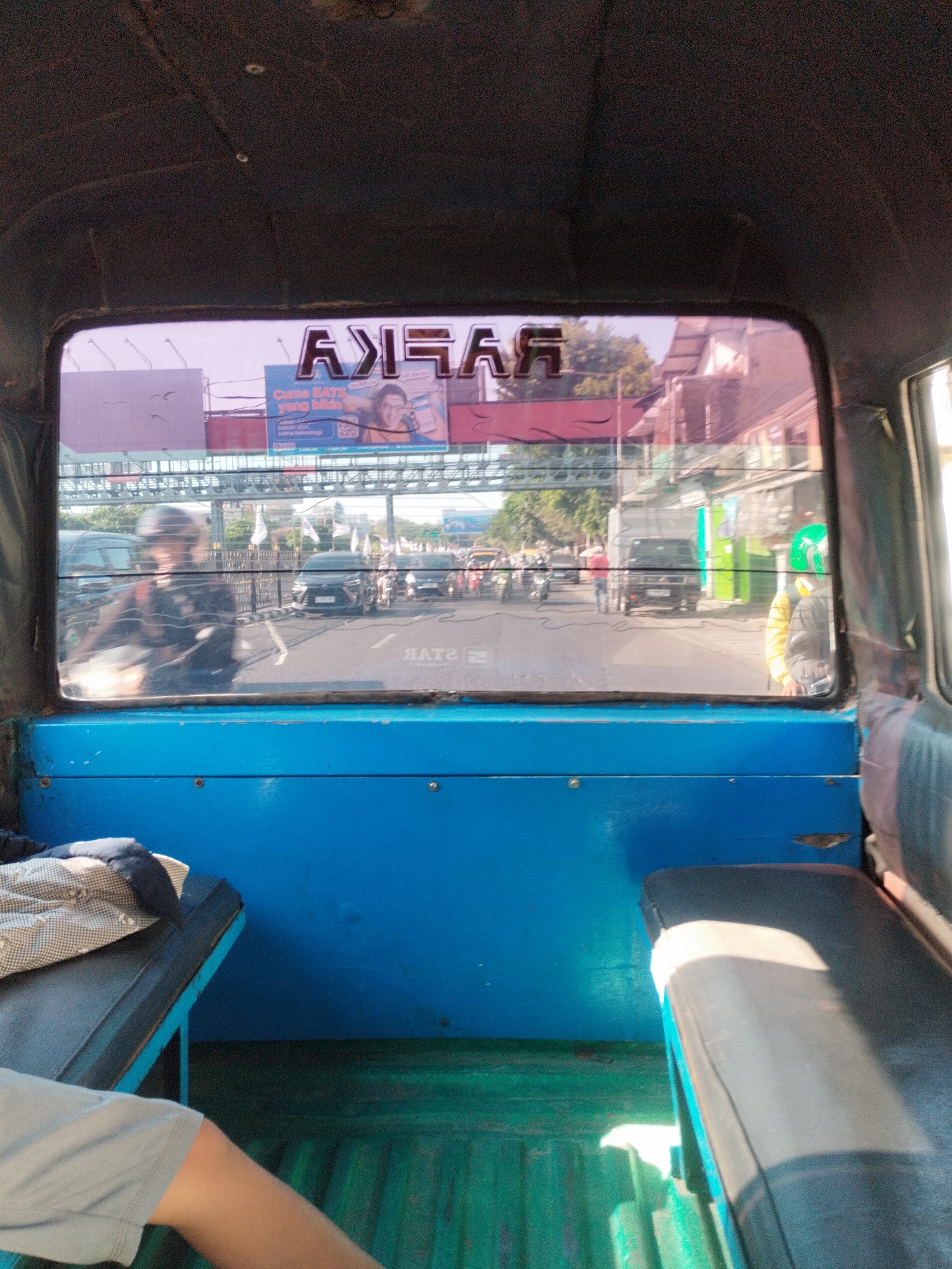 The image depicts a view from inside a vehicle, likely an open-air transport such as a bemo or pick-up truck, looking through the rear window at a street scene in Surabaya, Indonesia. The interior of the vehicle is characterized by blue painted panels and green ribbed flooring. Two dark-colored seats are visible, one with a patterned cushion.
Outside the vehicle, the street is busy with traffic. Several motorcycles and cars are present, moving in the same direction. A pedestrian in a red shirt is visible on the right side of the road, and another individual wearing a yellow jacket and green helmet is seen near the right edge of the frame. A large billboard with text that appears to be in Indonesian is situated above the street, featuring an image of a person holding a phone. The text on the billboard includes the words "Cuma EATS yang bikin" and "bukan sihir, cuma-teknologi." Reflected on the rear window of the vehicle is text that reads "ЯAT2" in reverse, likely from another sign. The overall environment suggests a daytime scene in a moderately developed urban area of Surabaya, characterized by typical Indonesian street traffic and advertising.