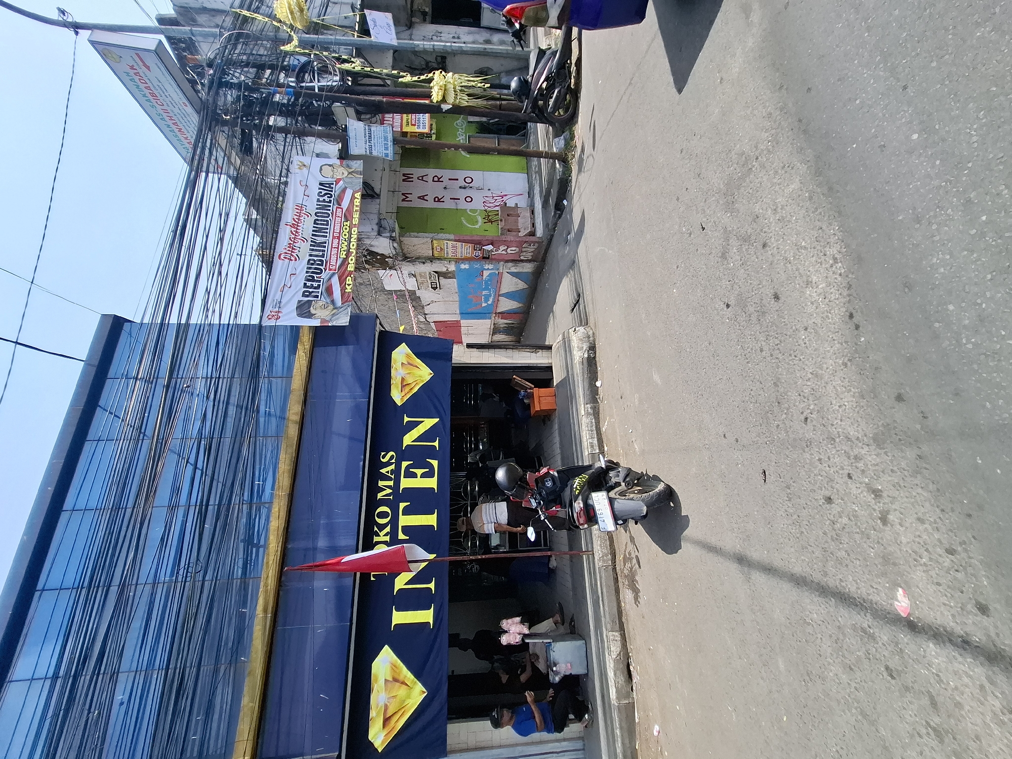 In the city of Cibadak, Indonesia, a street scene unfolds under a bright, clear sky. The foreground features an asphalt road with scattered debris. A black motorcycle, license plate F 4169 UCE, is parked on the right side of the road, casting a shadow. To the left, a sidewalk borders a building with a dark blue facade and a yellow awning. The building displays a prominent sign with large yellow lettering that reads "TOKO MAS INTEN," indicating a gold shop. Red and white Indonesian flags are displayed on poles in front of the shop. Several individuals are visible on the sidewalk in front of the gold shop, some appearing to be customers and others possibly employees. One person in a blue shirt sits on a stool, while others stand or are engaged in conversation. In the background, a white banner with red and blue graphics, celebrating "DIRGAHAYU REPUBLIK INDONESIA," is visible. Adjacent buildings display various signs and posters, including one with "MARIO" in large green lettering, suggesting commercial establishments. Numerous electrical wires crisscross overhead, a common sight in urban Indonesian environments. The overall atmosphere is that of a typical bustling street in a smaller Indonesian city, with a mix of commercial activity and daily life.