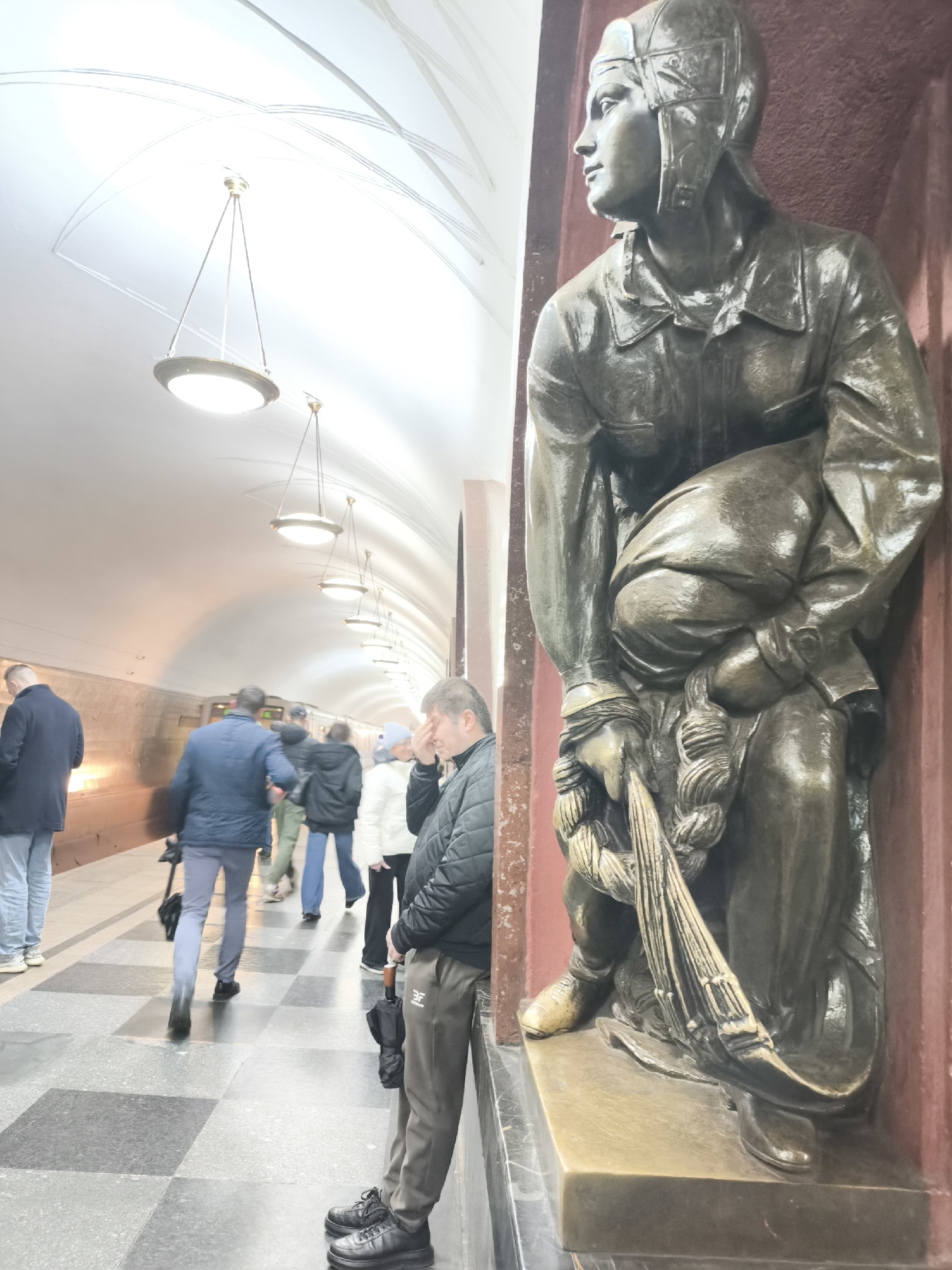 The image displays a bustling subway station platform, distinguished by a large bronze statue on the right and an arched, brightly lit ceiling.
The main subject on the right is a meticulously detailed bronze statue, likely depicting a pilot or aviator. The figure wears a distinctive helmet, a jacket, and is in a crouching or seated posture, gazing intently to the left. The statue holds braided ropes or cables in its hands and is set against a deep red wall.
To the left, the subway platform extends into the distance, illuminated by a series of elegant, circular hanging lights that punctuate the arched white ceiling, which also features subtle decorative lines. Numerous commuters are visible, mostly blurred due to motion as they walk along the platform. They are dressed in jackets, and some carry umbrellas. In the immediate foreground on the left, a man leans against the wall, holding a closed black umbrella. He has his hand to his forehead, suggesting weariness or pensiveness. A faint two-letter logo, possibly "BF", is visible on his trousers.
The platform floor features a checkered pattern of light and dark grey squares. In the background, a subway train is partially visible, indicating active service. The architectural style of the station, with its grand ceiling and detailed statue, points to a historically significant metro system, possibly in a city renowned for ornate stations. The artificial lighting suggests it could be any time from day to evening, while the clothing and umbrellas hint at cooler or rainy weather.