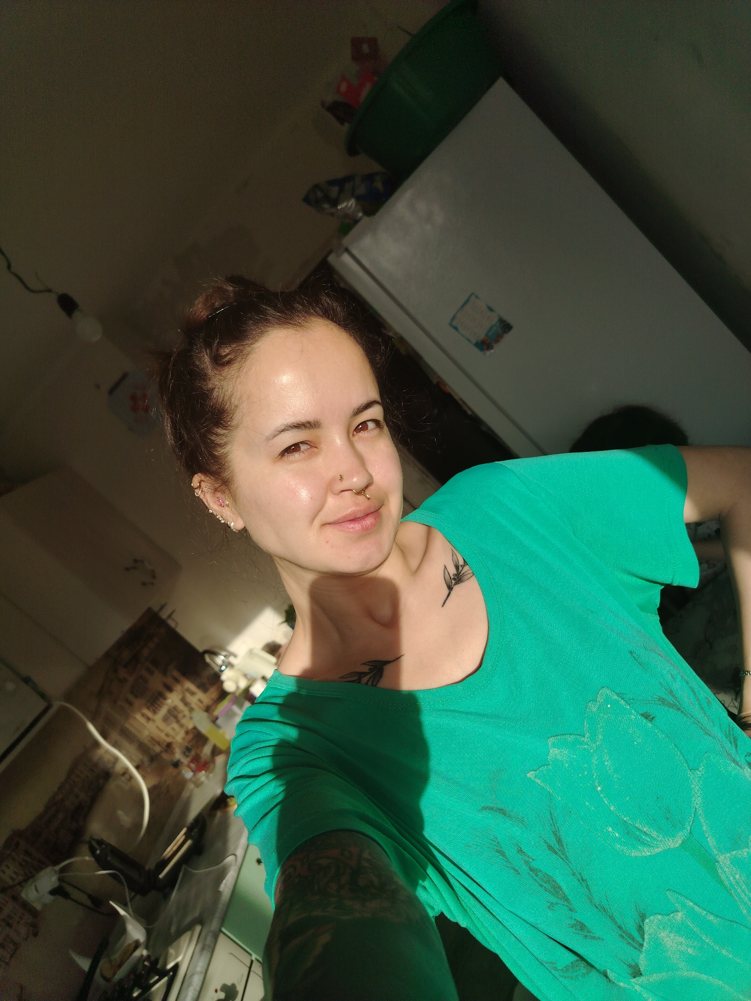 A woman is posing for a selfie in what appears to be a kitchen. She is wearing a green t-shirt with a floral design, has her hair up in a bun, and has multiple piercings in her ear and nose. She has tattoos on her chest and arm. The kitchen appears somewhat cluttered. There is a white refrigerator in the background, along with various kitchen appliances and items. The location is identified as being in Noviy, Russia.