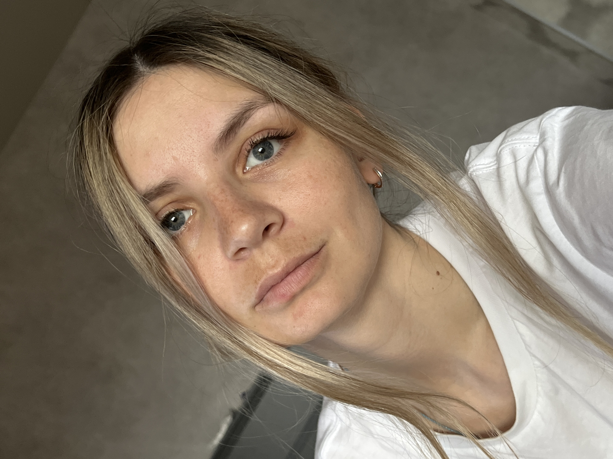 A fair-skinned woman with long blonde hair is shown in a close-up selfie. Her eyes are blue, and she has freckles on her face. She wears a white t-shirt and a small hoop earring in her left ear. The background appears to be a gray wall or surface. There is no indication of location in Chaikovsky, Russia, or any other city.