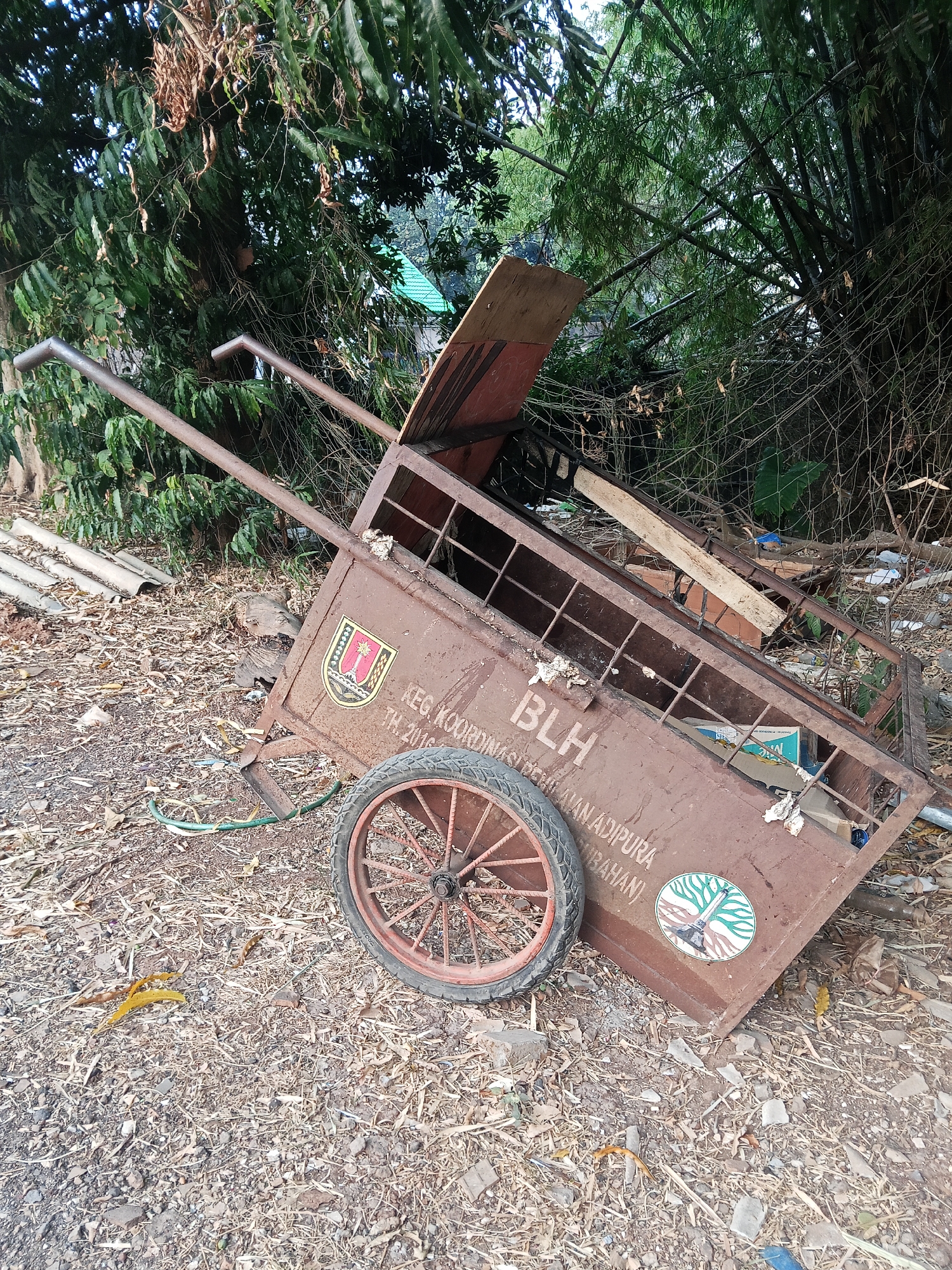 An outdoor, ground-level, eye-level, full shot captures a rusty, brown, two-wheeled cart, or pushcart, situated in a cluttered, outdoor environment. The cart is tilted and appears to be empty, except for some debris visible inside. The cart has two metal handles at the top for pushing and steering. A single, spoked wheel with a worn tire is attached to the rear of the cart. The cart has text and emblems on its side. One emblem is a crest with a tower and what appears to be a sheaf of wheat. Below the crest, text reads "KEG. KOORDINASI PENGELOLAAN ADIPURA." Another emblem, circular in shape, depicts a stylized tree with roots and a central figure. The cart is surrounded by a mixture of dry leaves, twigs, small rocks, and some white and blue plastic debris. In the background, there is dense green foliage, including bamboo trees, and a glimpse of a green roof. The scene is indicative of a neglected or disused area, potentially on the outskirts of a residential or industrial zone. The cart bears markings that suggest it may be associated with waste management or public services in Semarang, Indonesia, specifically referencing "PENGELOLAAN ADIPURA," a program related to environmental cleanliness and city beautification.