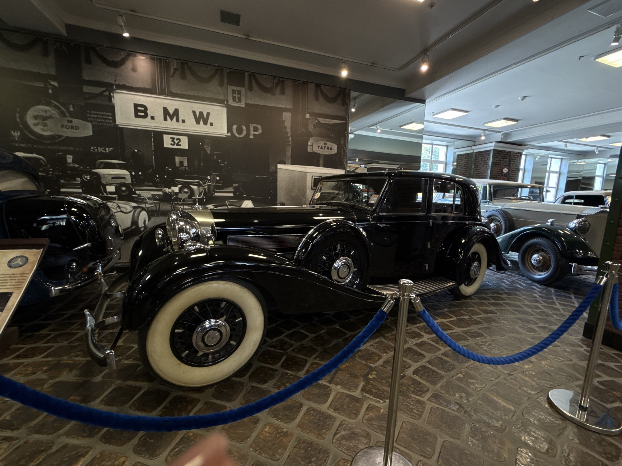 The image depicts the interior of a museum exhibit featuring vintage automobiles. In the foreground, a black vintage car with white-walled tires and chrome accents is prominently displayed. A blue velvet rope barrier, supported by chrome stanchions, separates the viewer from the car. Behind this car, other vintage vehicles, including a light-colored car, are visible. The background features a large black and white mural of historical automobiles and signage, including "B. M. W." and "FORD". The exhibit is situated on a stone-paved floor and illuminated by overhead lighting. The location is identified as the city of Arkhangel'skoye, Russia, likely within a museum setting.