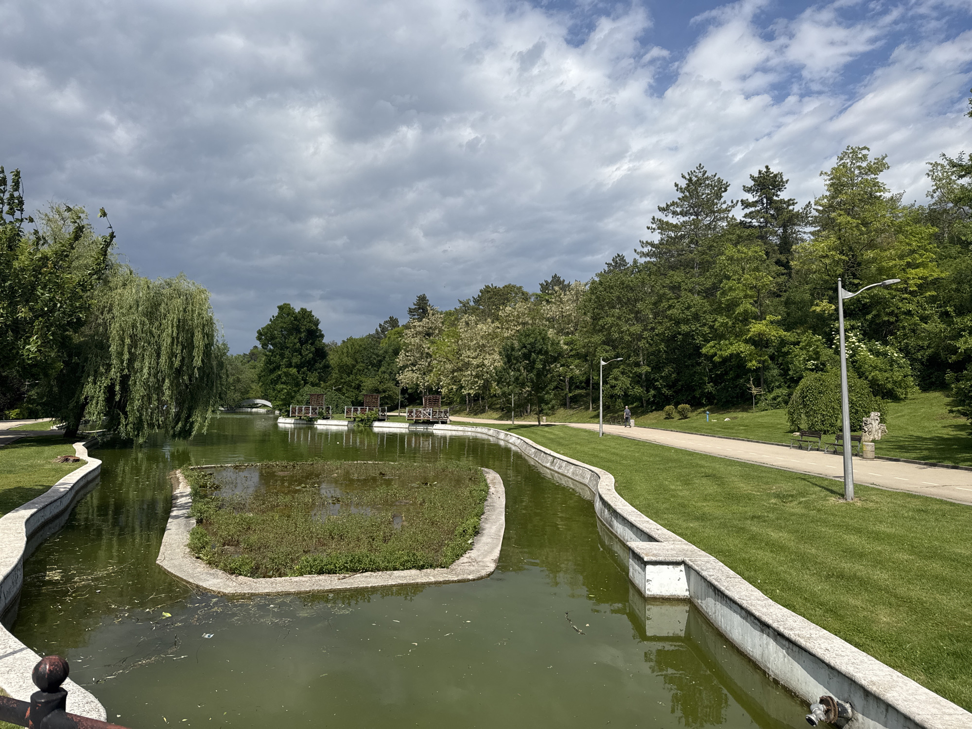 The image depicts a park in Dobrich, Bulgaria, featuring a waterway with concrete edging. The water appears green and contains an island covered in vegetation. A weeping willow tree is on the left. A pedestrian walkway runs alongside the water, and a person is walking in the distance. There are trees and other greenery in the background. The sky is partially cloudy.