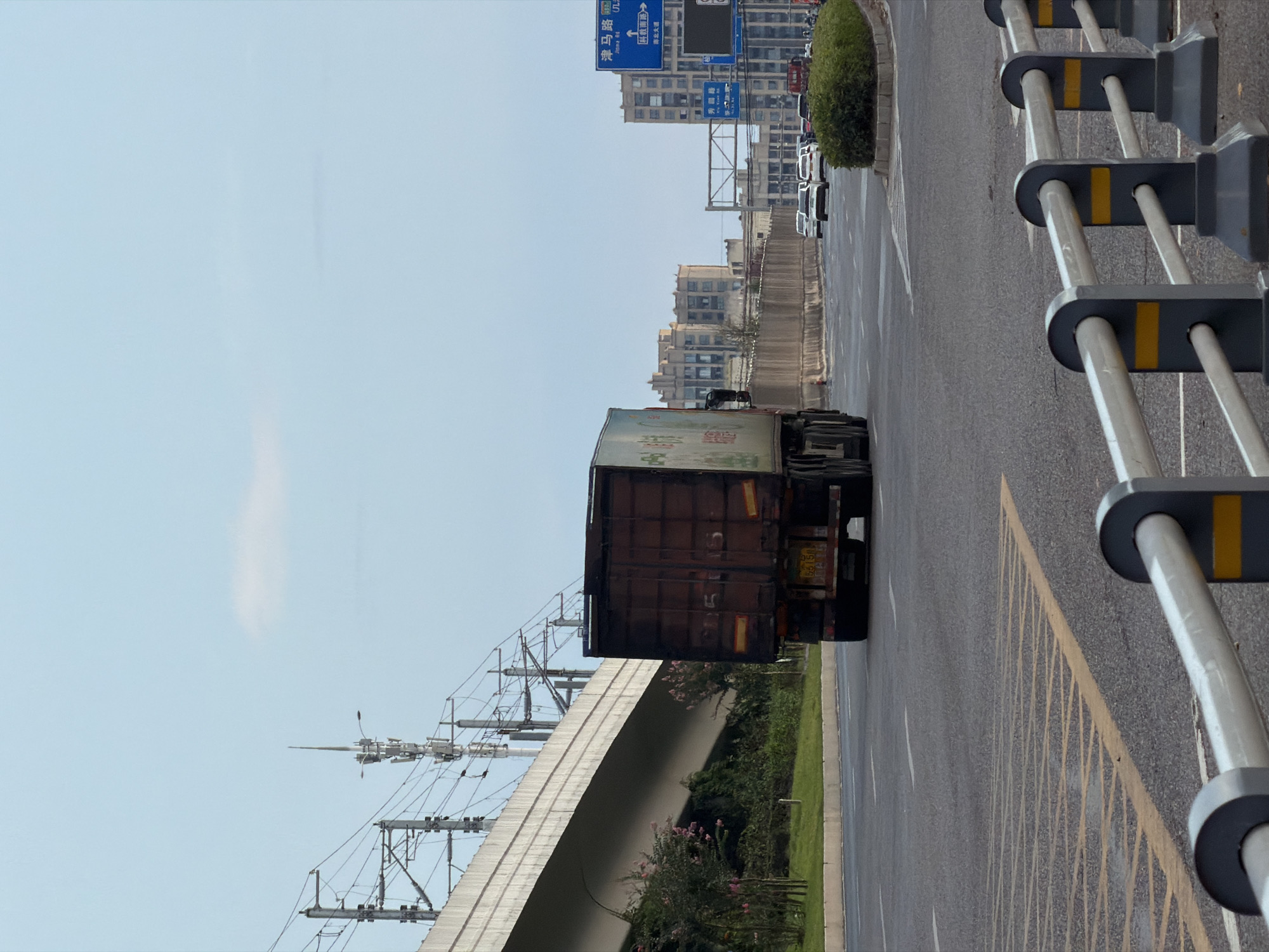 A large, dark-colored semi-trailer truck drives down a multi-lane highway in Jiangjin District, Chongqing, China, under a clear, pale blue sky with a few wispy clouds. The truck is traveling away from the camera, and its cargo container is dark brown with some faded markings. The license plate is visible and appears to be yellow with black Chinese characters and numbers.
To the left of the highway, a concrete overpass with overhead electrical lines for a railway runs parallel to the road. Green vegetation and scattered pink flowering bushes are visible on the embankment beneath the overpass.
On the right side of the highway, traffic signs are posted on blue poles. One sign, with Chinese characters and some English text, indicates directions. Further down the road, several cars are driving in the same direction as the truck. Beyond the highway, a cityscape of multi-story buildings can be seen. The highway is bordered by a median with manicured bushes and a metal guardrail. The road surface is asphalt, with yellow lane markings visible. The overall scene depicts a typical urban transportation artery.