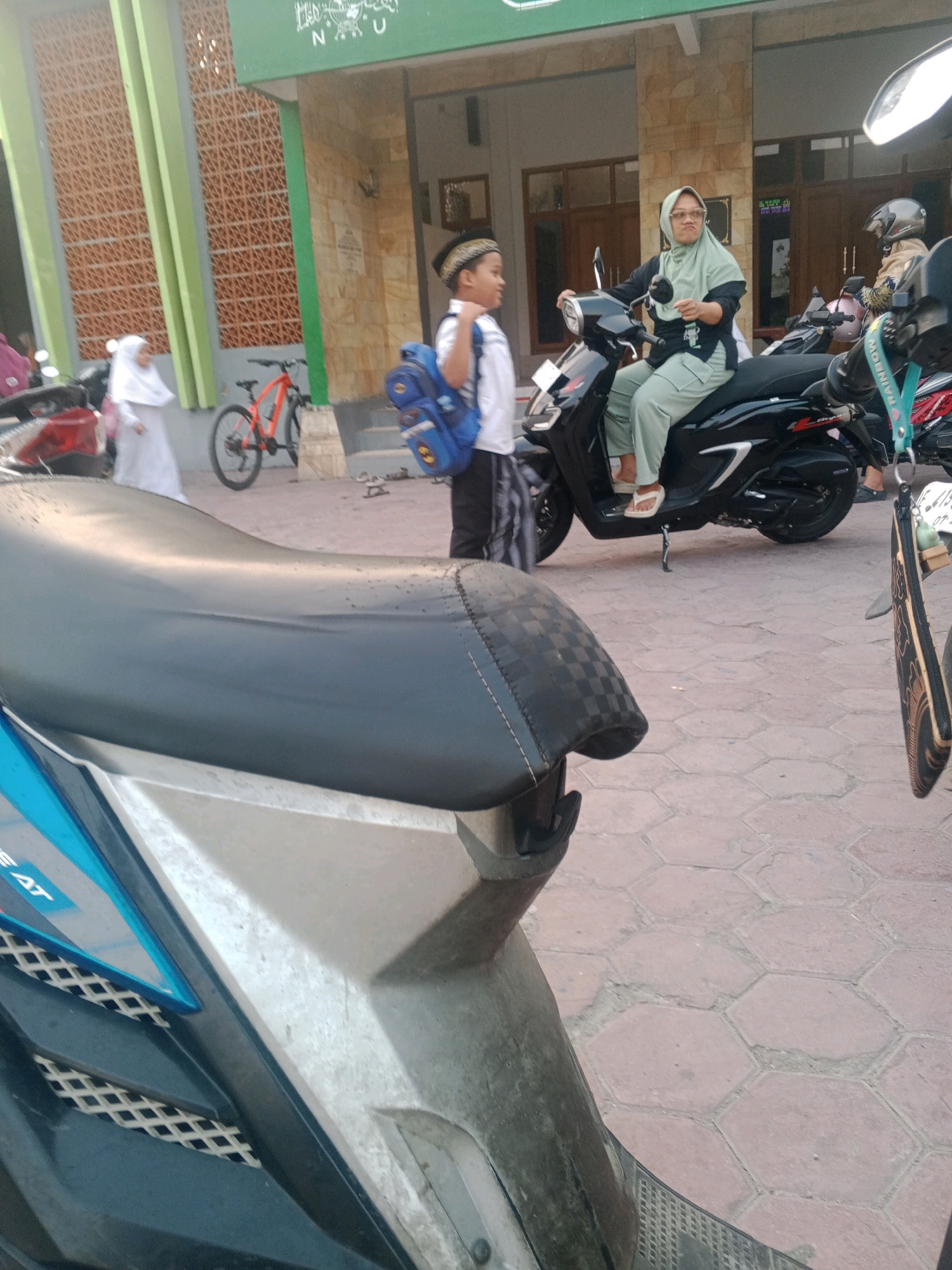 A wide-angle, eye-level shot captures an outdoor scene in Lembah, Indonesia, likely near a place of worship given the architecture. In the foreground, the dark seat of a parked motorcycle occupies the lower left quadrant. To the right of the motorcycle, a woman wearing a pale green hijab and matching pants sits on a black scooter, looking towards the left of the frame with a slightly pursed expression. Her left hand rests on the scooter's handlebar. Beside her, a young boy in a white shirt, black and white patterned pants, and a white skullcap stands facing away from the camera, carrying a blue backpack. Behind them, a red bicycle is parked. In the background, a building with light-colored stone and green accents is visible, with a wide entryway. Other motorcycles and people are also present, suggesting a public space. The ground is paved with reddish-brown hexagonal tiles. The lighting suggests it is daytime, with the sun casting some shadows.