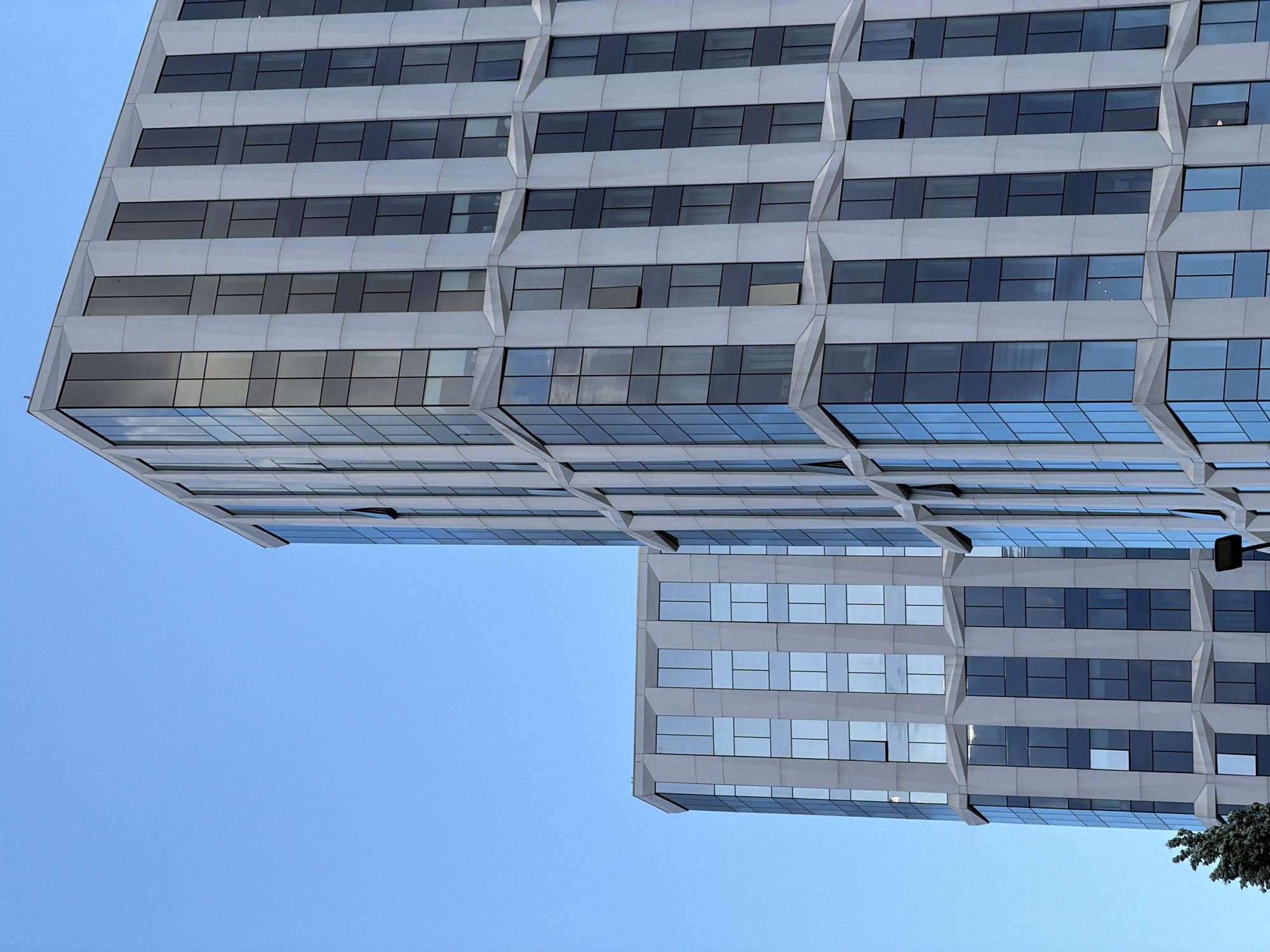 A low-angle shot reveals the facades of two adjacent high-rise buildings against a clear blue sky. The buildings feature a repetitive pattern of horizontal bands and inset windows, creating a textured and geometric appearance. The color palette is limited to the pale grey of the building's facade and the dark reflections in the glass windows. The composition of the shot focuses on the architectural details and the relationship between the buildings, rather than providing a wide contextual view. The city is unknown.