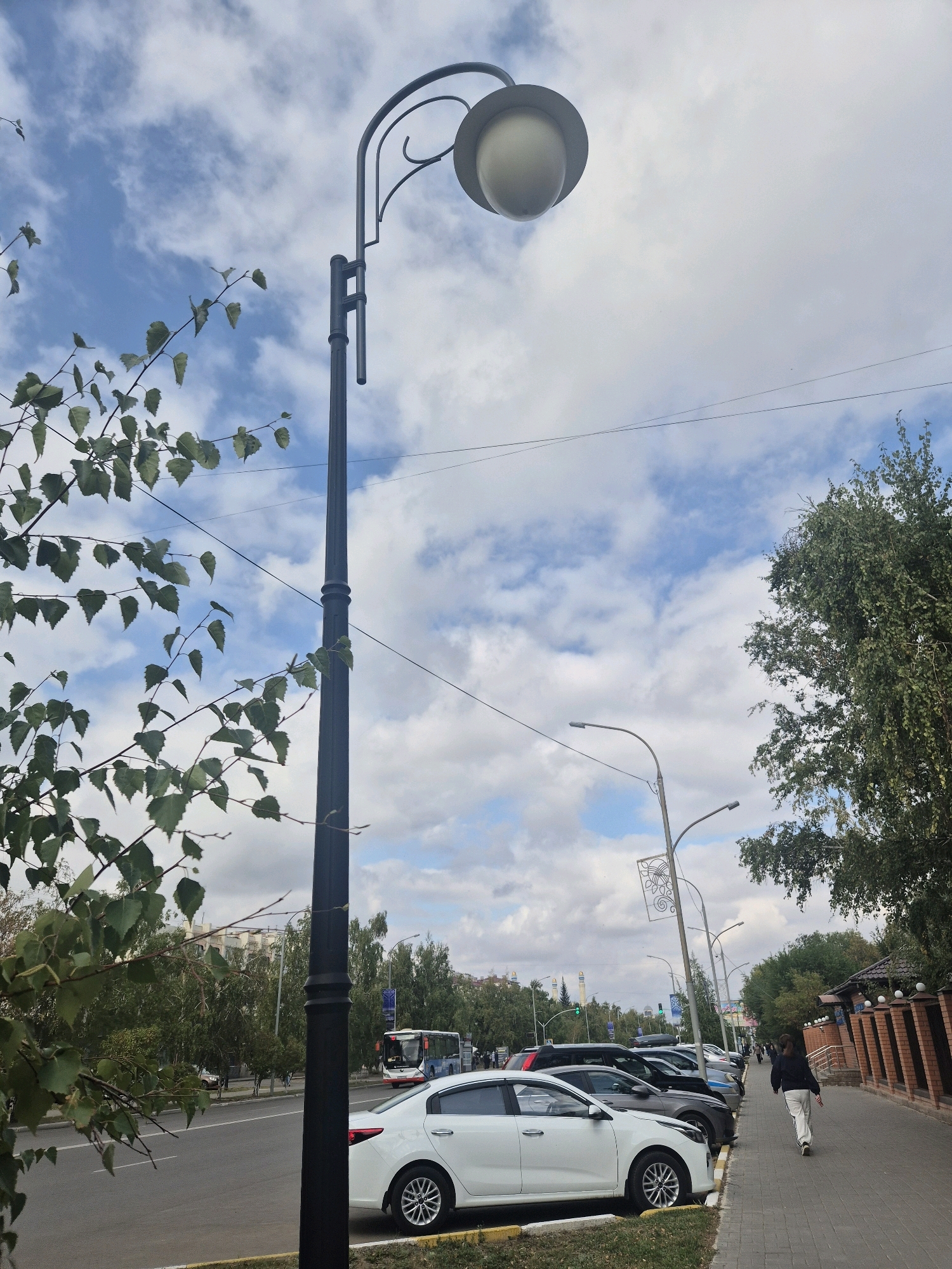 This is a street-level, eye-level shot taken on a cloudy day. In the foreground, a tall black lamppost stands to the left, its curved arm supporting a round, cream-colored lamp. To the left of the lamppost, branches of a green-leaved tree extend into the frame from the upper left corner. The street is visible with several parked cars, including a white sedan in the foreground. A bus is seen driving in the distance on the road. A paved sidewalk runs along the right side of the street, with a person in dark clothing and white pants walking away from the camera. A brick fence with decorative elements lines the right edge of the sidewalk, beyond which is dense green foliage. The background shows a mix of trees and buildings, with a blue sky dotted with white clouds overhead. The scene appears to be a typical urban street in Kokshetau, Kazakhstan, likely a residential or commercial area.