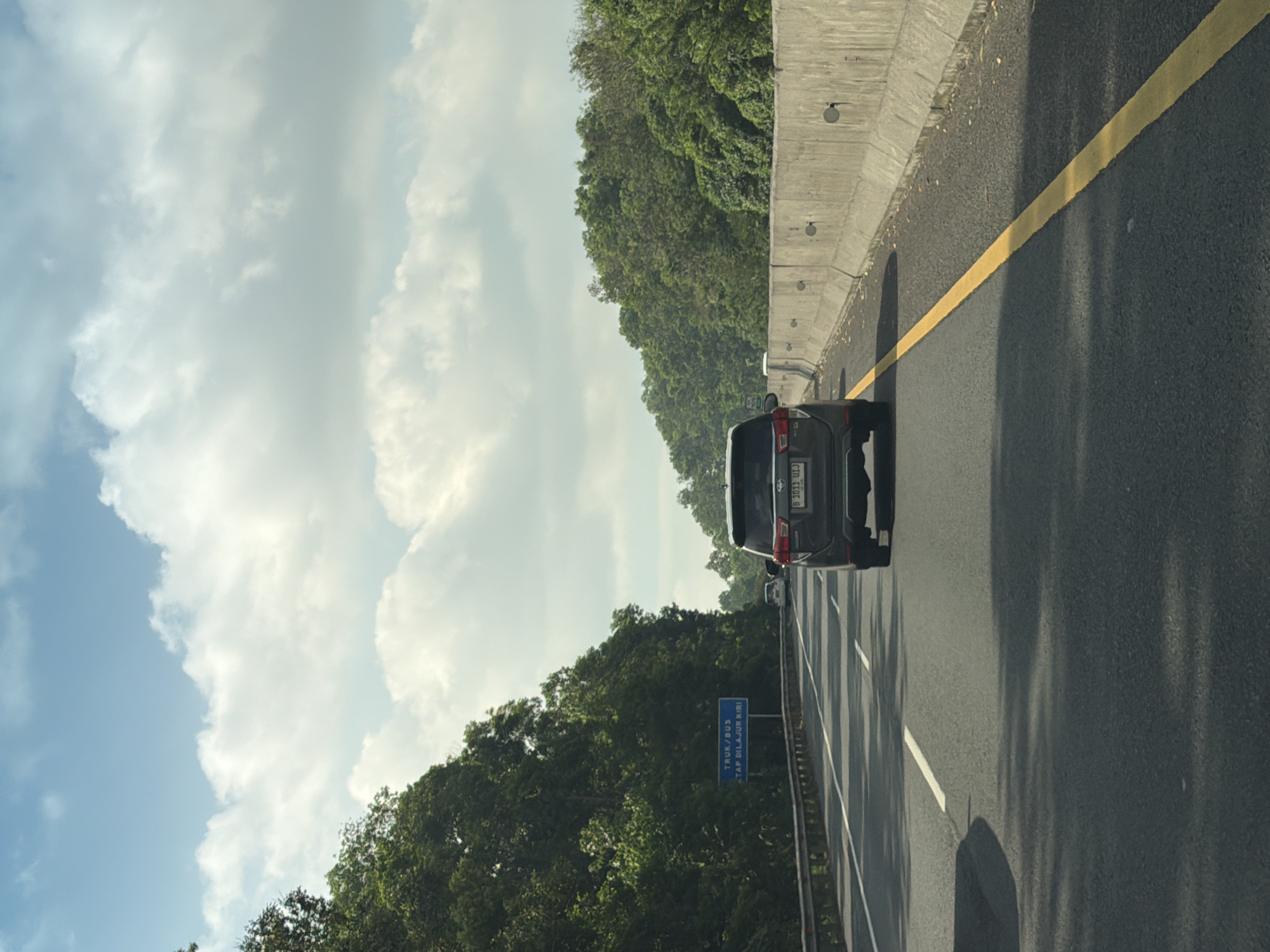 A dark-colored SUV is driving on an asphalt highway in Mandalasari, Indonesia. The SUV is in the right lane, and there is another vehicle in the left lane further ahead. The highway is lined with lush green trees on both sides. A concrete barrier runs along the right side of the road, and a yellow line marks the edge of the lane. The sky is partly cloudy with patches of blue sky visible. A blue sign on the left side of the highway reads "TRUK/BUS TETAP DILALUI KIRI." The overall scene depicts a typical highway drive in a tropical environment.