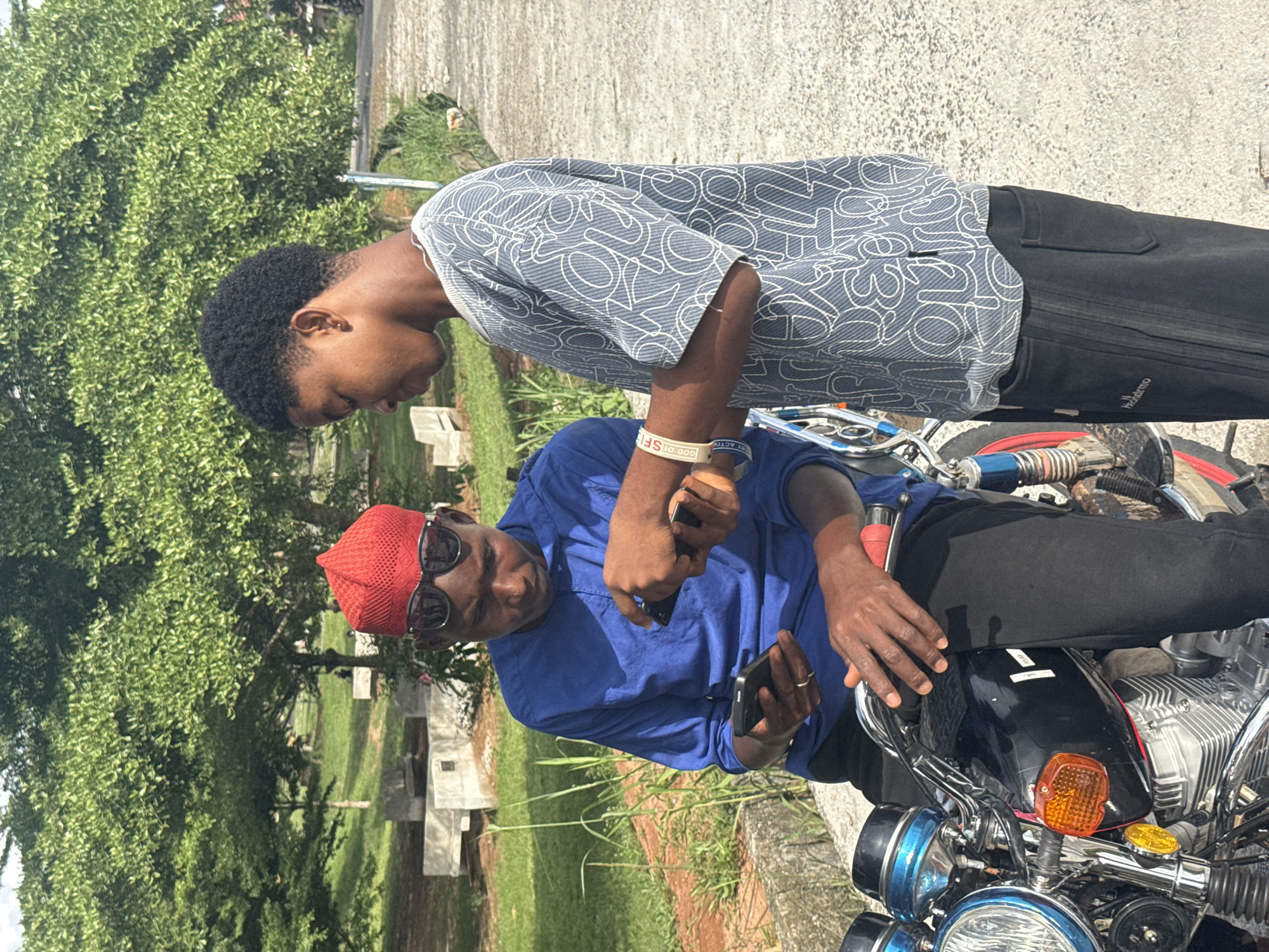 Two men are positioned on or near a motorcycle. One man, wearing a blue patterned shirt and black pants, is standing and appears to be looking down. The other man is seated on the motorcycle, wearing a blue shirt, black pants, sunglasses, and a red hat. He is holding a phone. The motorcycle has a blue headlight housing, a black front fender, and red accents on the wheel rims. The setting appears to be outdoors, with trees, grass, and a concrete path visible. The location is identified as Ihiagwa, Nigeria.