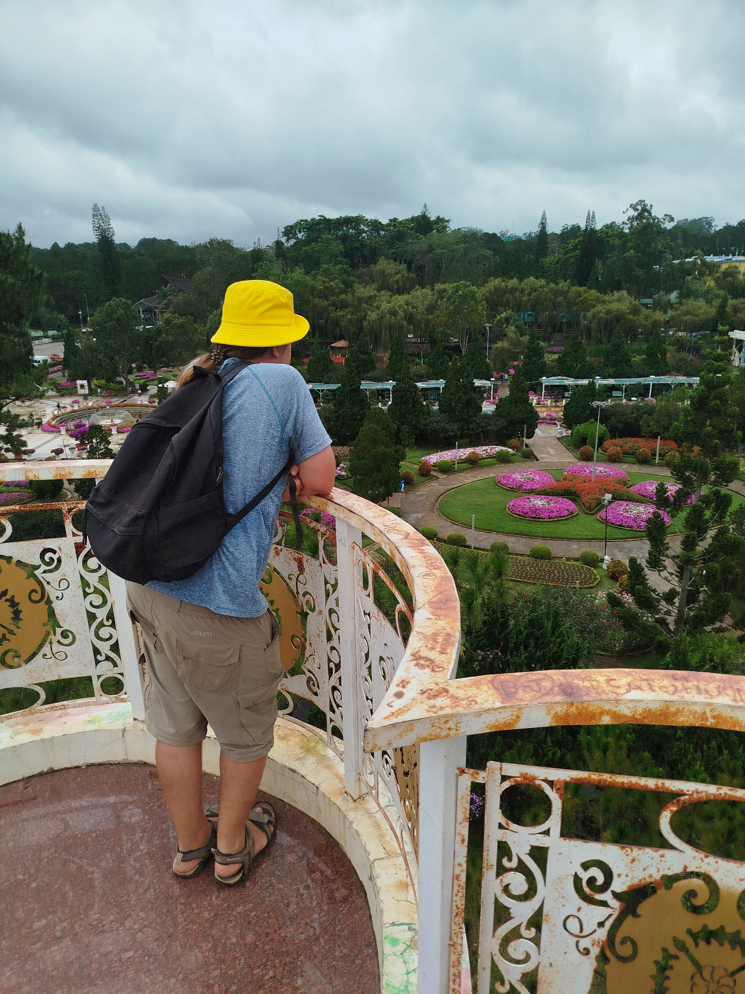 A person wearing a yellow bucket hat, a blue t-shirt, khaki cargo shorts, and brown sandals with a black backpack stands on a circular balcony overlooking a garden. The balcony railing is white with ornamental cutouts and has areas of rust. The person is looking out at the garden, which features manicured flower beds with pink and red flowers, green lawns, trees, and pathways. The sky is overcast. The scene appears to be located in a park or botanical garden.