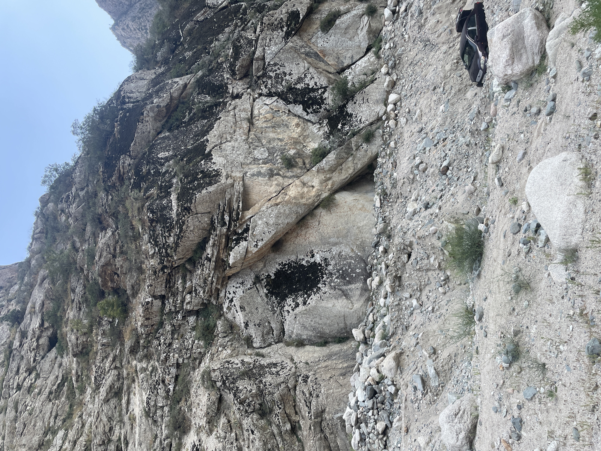 The image depicts a dark-colored sedan partially submerged in a debris-filled ravine on a steep, rocky mountainside. The car's trunk is open. The surrounding environment is characterized by a dry, rocky terrain with scattered stones and sparse vegetation. The mountainside features exposed rock faces with patches of dark moss and lichen. The sky above is clear and pale blue. The scene appears to be located in a remote, mountainous area, consistent with the description of Khodzha-Obigarm, Tajikistan, known for its natural hot springs and rugged landscape.