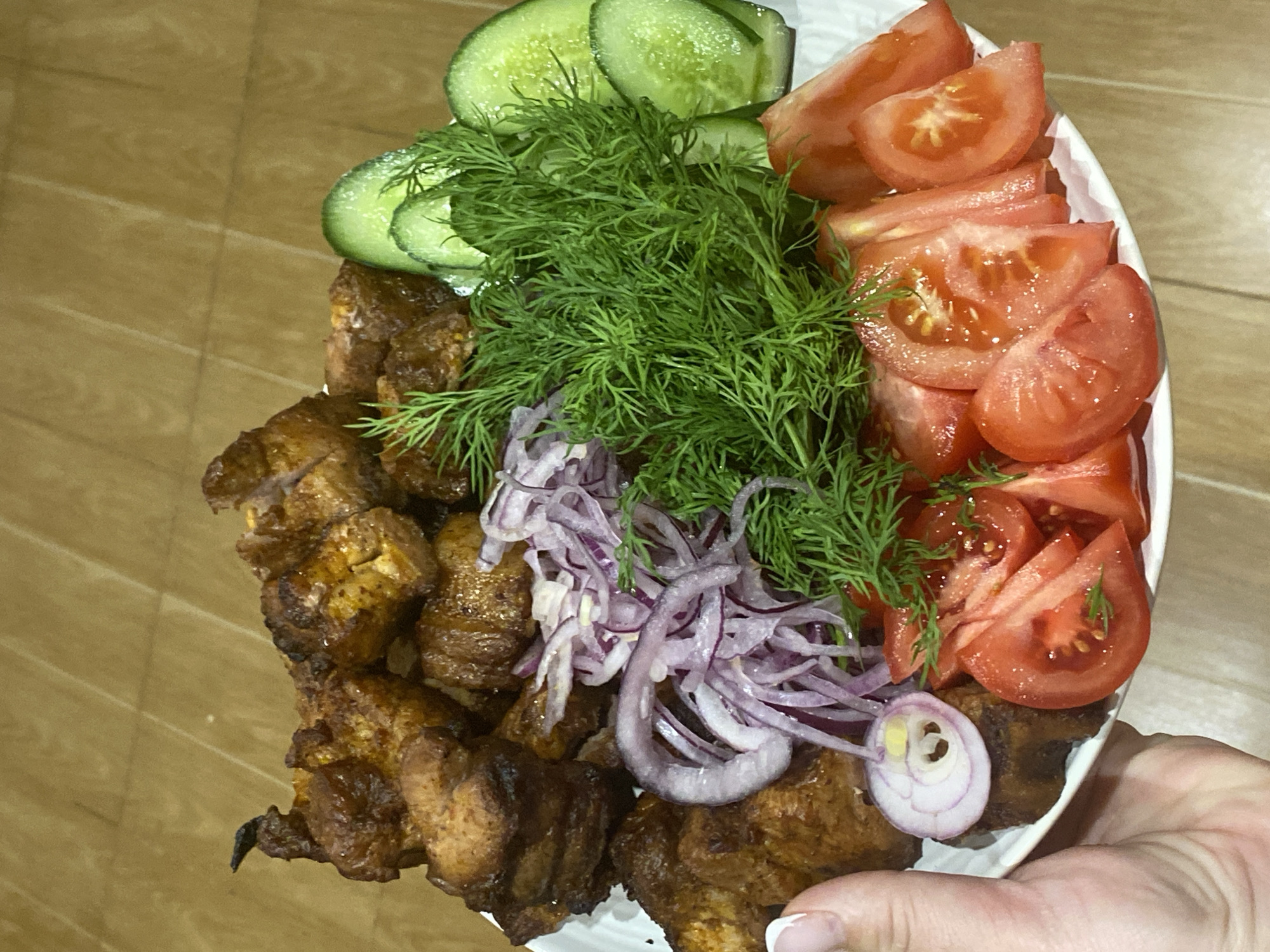 This close-up, high-angle shot captures a white oval plate laden with a traditional meal, held by a person's hand in the bottom right corner. The main subject is a generous portion of shish kebab, consisting of cubed, grilled meat pieces, dark brown and slightly charred, occupying the left side of the plate.
Accompanying the meat is a fresh salad arrangement. On the top left, several bright green cucumber slices are visible. Directly beside the cucumbers and extending across the middle is a large mound of fresh, vibrant green dill. To the right, numerous wedges of ripe red tomatoes are neatly arranged. A scattering of thinly sliced purple-red onions is placed over the shish kebab and nestled among the dill.
The food appears freshly prepared, with the vegetables looking crisp and the meat having a well-cooked appearance. The plate is presented against a light brown, wooden background, possibly a table or floor. A hand with manicured fingernails is visible supporting the plate from underneath. The composition is bright and colorful, showcasing the meal in an appealing manner. There is no visible text in the image. This type of meal, shish kebab with fresh vegetables and herbs, is a common dish found in Russia and the Caucasus region, consistent with the Sochi, Russia location context.