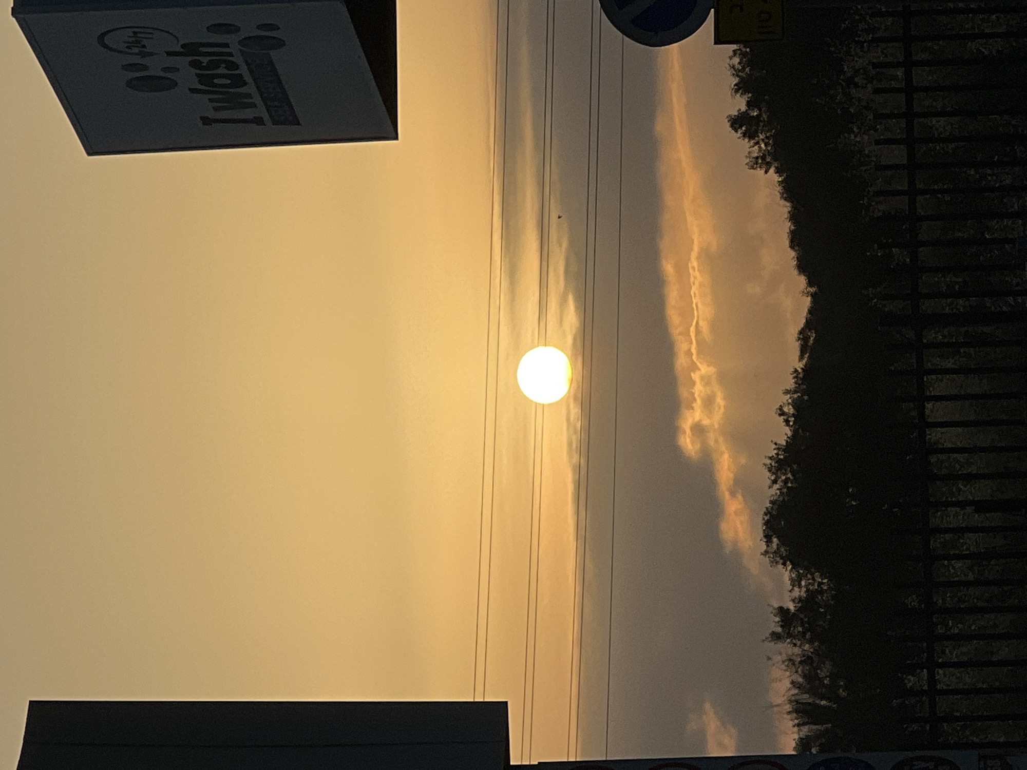 The image captures a striking sunset or sunrise scene, dominated by a brilliant, almost white circular sun positioned centrally in the frame. The sky transitions from a vivid golden-orange hue around the sun to softer yellows and grays towards the edges.
Several thin, dark parallel lines, likely utility wires, stretch horizontally across the sky, passing behind the sun. A small, dark, indistinct shape, possibly a bird, is visible in the upper center of the sky.
In the upper left, a dark, rectangular sign is visible, displaying white text. "24/7" is printed at the top, followed by the larger, more prominent "I wash". A smaller, less legible line of text, possibly "SELF SERVICE", is beneath "I wash". The lower left corner shows the top edge of another dark, silhouetted structure.
Along the right side and bottom right, a dark, silhouetted treeline or dense foliage provides a stark contrast against the illuminated sky and a elongated, golden-orange cloud. A dark, grid-like or fence-like structure is faintly discernible behind the treeline on the far right. The overall atmosphere suggests a peaceful moment during dawn or dusk in Beer Yaakov, Israel.