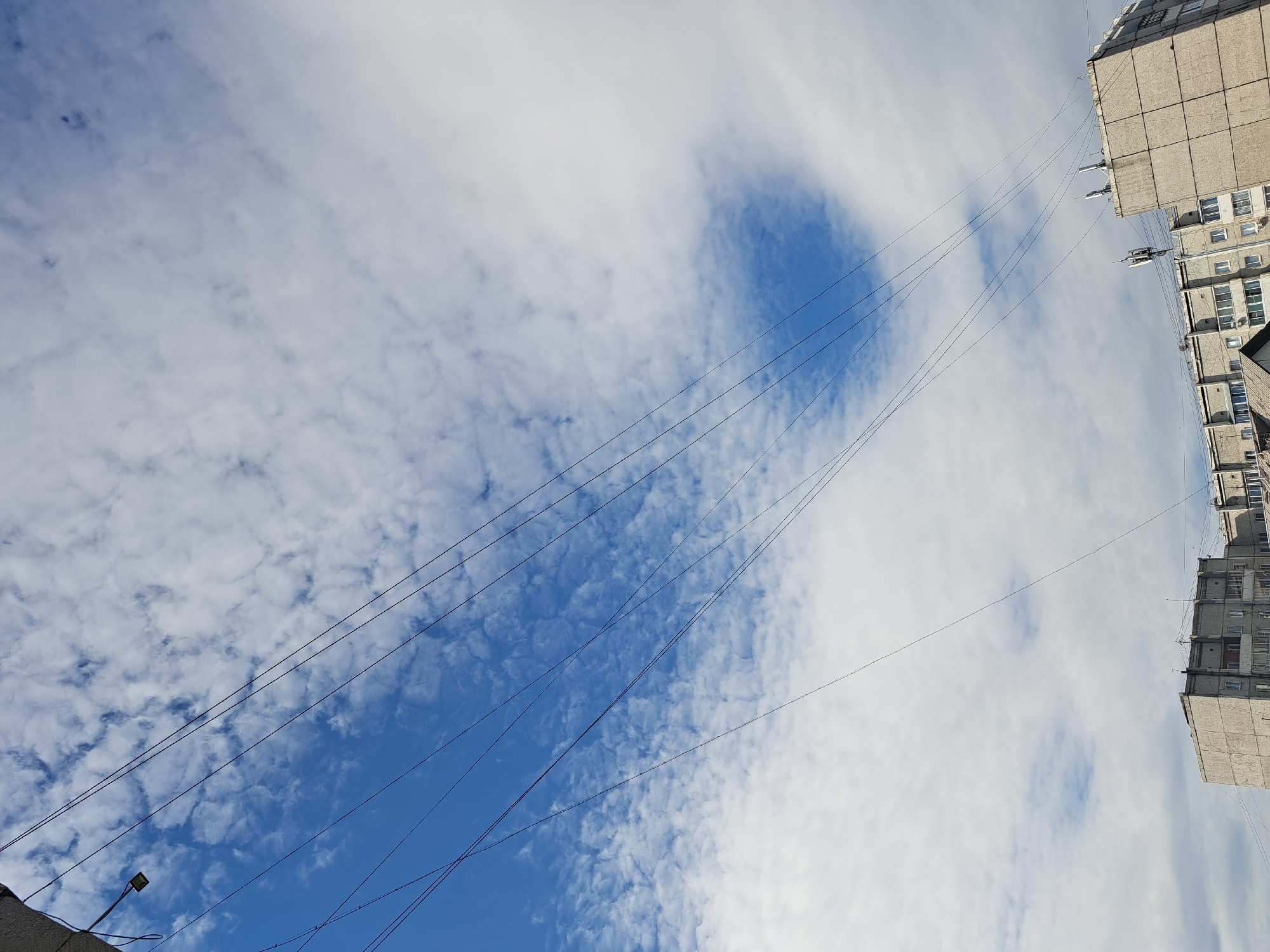 This is a medium-angle, outdoor, eye-level shot of a cloudy sky above a residential building in Irkutsk, Russia. The image is dominated by the sky, which features a mixture of white, fluffy clouds and patches of clear, bright blue. Multiple thin black wires stretch diagonally across the frame from the top left to the bottom right, suggesting they are power or communication lines. In the lower portion of the image, the top sections of two monolithic, concrete apartment buildings are visible. These buildings appear to be typical Soviet-era architecture, with multiple stories and rows of windows. One building on the right has several antennae or communication dishes mounted on its roof. There are no people visible in the frame. The overall impression is one of an urban, residential environment under a dynamic sky.