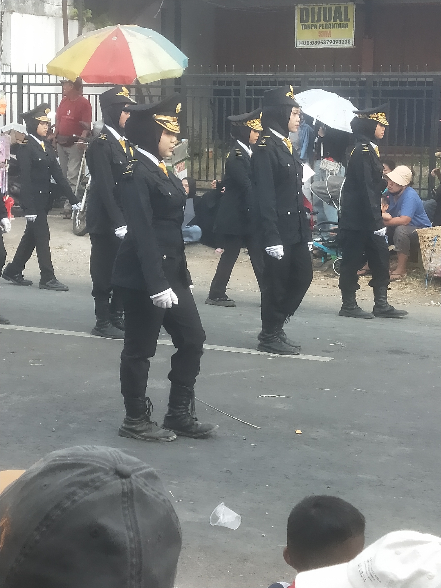 In Boja, Indonesia, a group of people in uniform are marching in a parade. Several individuals are wearing black uniforms with gold trim and hats, some are also wearing hijabs. One person in the foreground is wearing a black hat. In the background, there is a yellow sign that says "DIJUAL TANPA PERANTARA" and a phone number. There is also a colorful umbrella, a fence, and other people watching the parade. The ground is paved, and there is a white line on the road. The overall scene depicts a public event or celebration in a town setting.