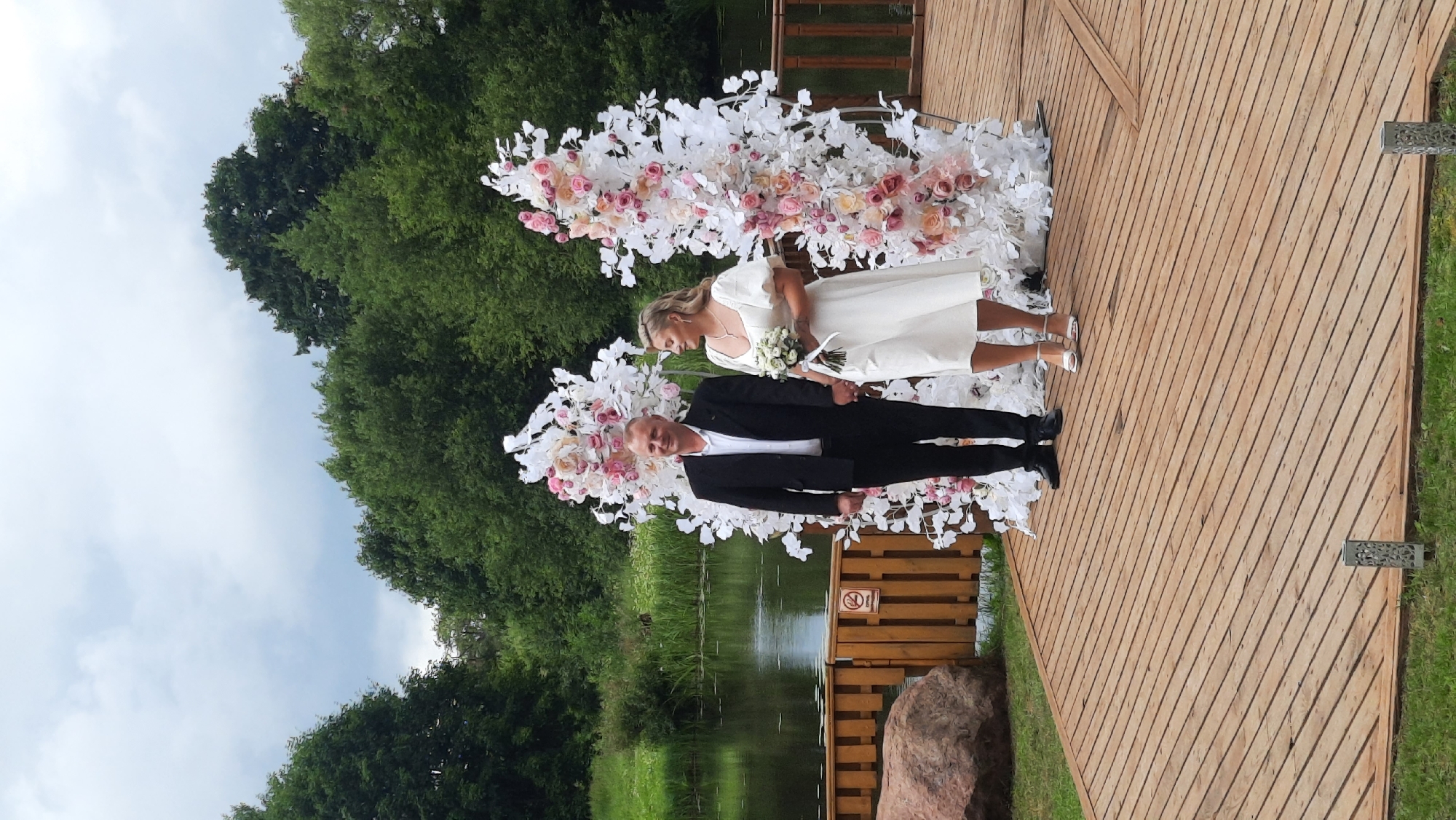 A wedding ceremony is taking place outdoors on a wooden deck in Жодзішкаўскі сельскі Савет, Belarus. A man in a black suit and a woman in a white dress are standing hand-in-hand in front of a floral wedding arch. The arch is decorated with white and pink flowers and branches. Behind them, a body of water is visible, with lush green trees lining the banks. The sky is overcast with clouds. The wooden deck is situated on a grassy area, with a large rock and a wooden fence to the left of the couple.