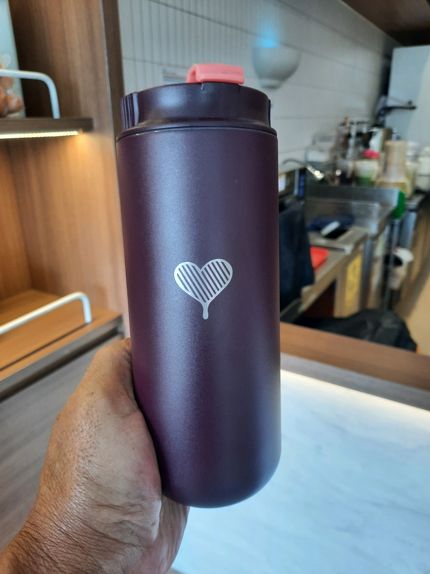 A person's hand is holding a dark purple thermos with a white heart logo. The thermos has a pink lid. In the background, there is a counter with various items, including bottles and dispensers. The scene appears to be indoors, possibly in a cafe or restaurant. The background suggests the location is East Jakarta, Indonesia, though no specific landmarks are visible.