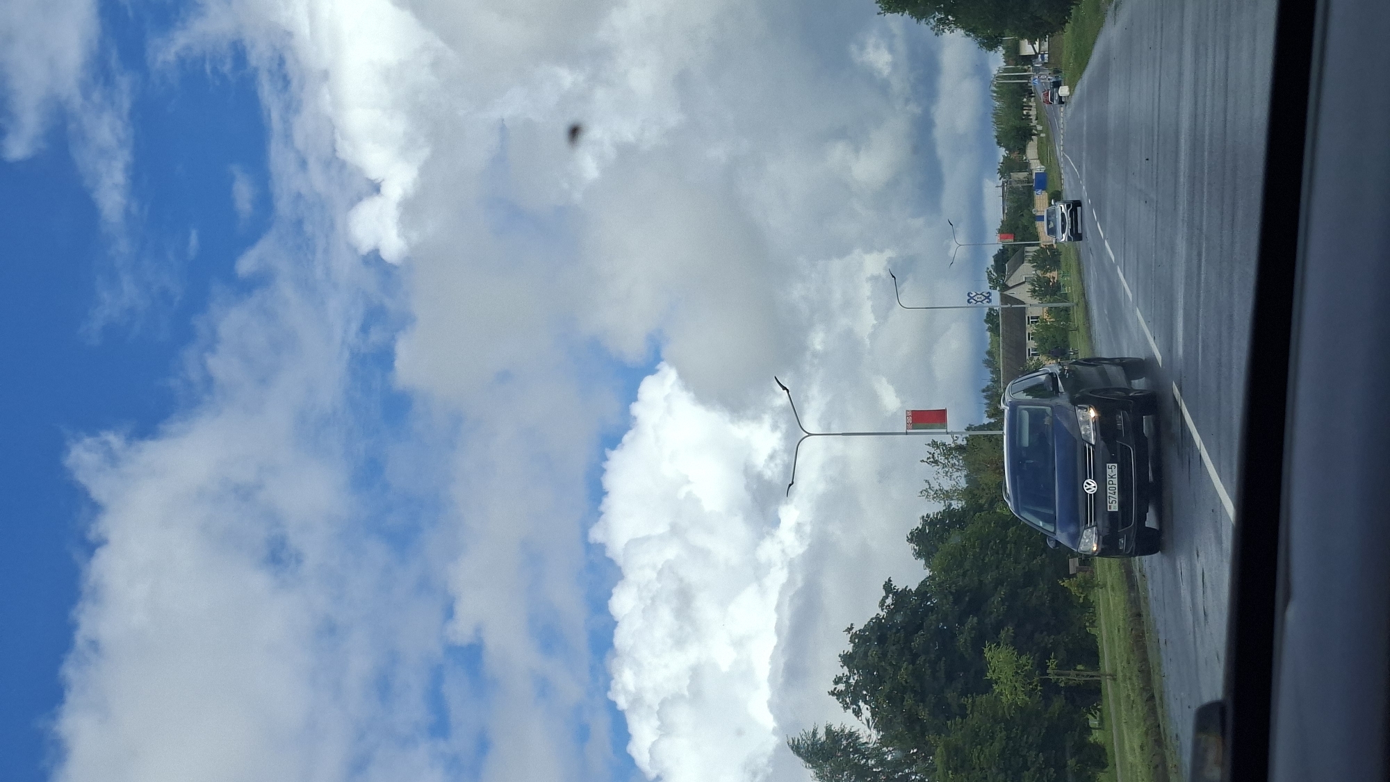 The image captures a view from inside a vehicle, looking out at a wet road in Myadzyel, Belarus. The sky is a dramatic mix of bright blue and large, puffy white clouds, with some darker, more ominous clouds also present. Along the roadside, there are streetlights with red banners hanging from them. Trees line the left side of the road, and a few houses and buildings are visible in the distance on the right.
Several cars are on the road. In the foreground, a dark Volkswagen Sharan is driving towards the viewer. Its license plate reads "5740PK-5". Further down the road, another white car is visible, also moving away from the viewer. In the far distance, other vehicles can be seen, suggesting normal traffic flow. The road surface appears wet, indicating recent rain. The overall impression is that of a cloudy but not necessarily rainy day, with a sense of movement and travel.