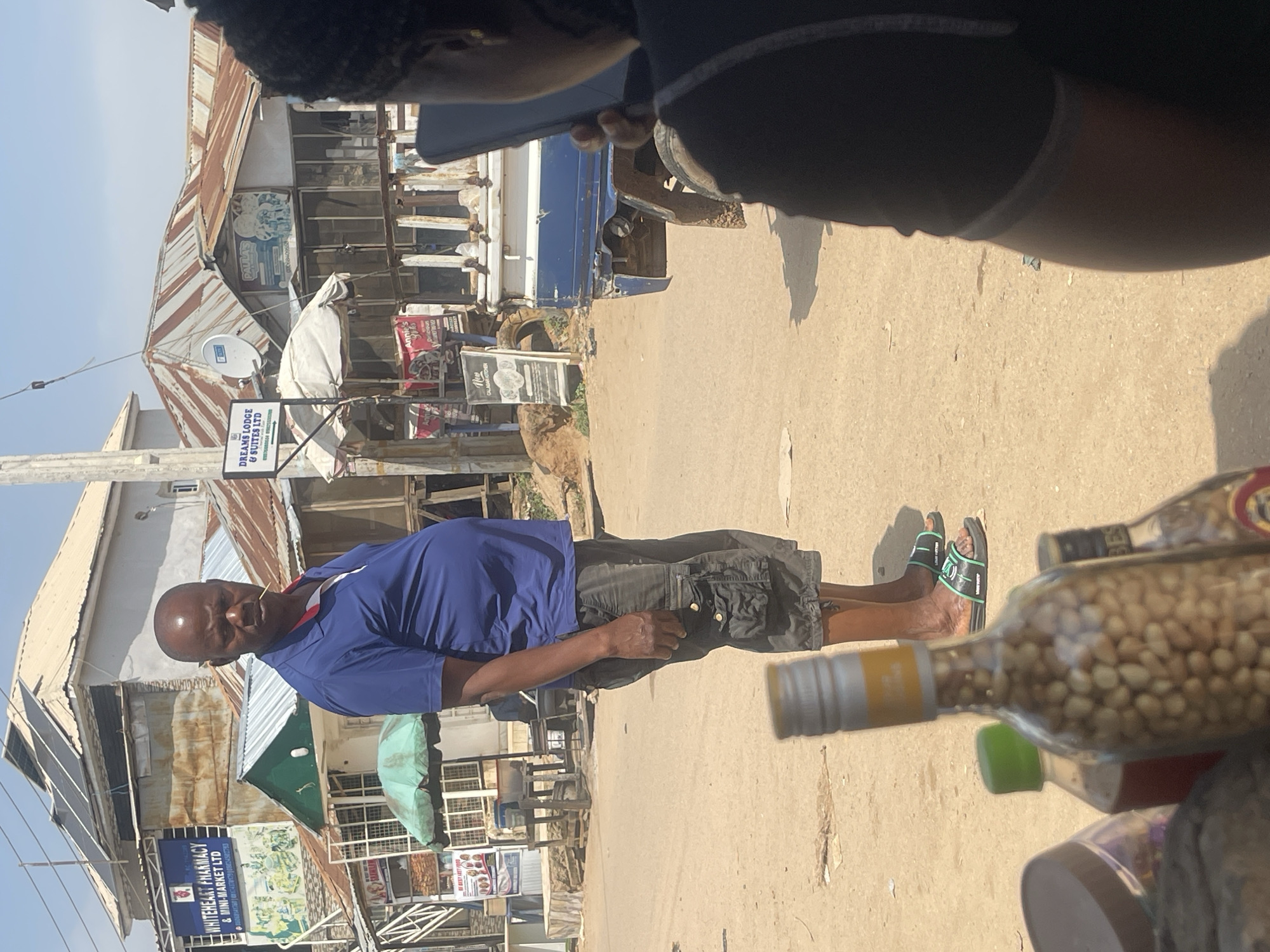 In Abuja, Nigeria, a man stands in an outdoor marketplace, facing left. He is wearing a blue polo shirt and dark cargo shorts. He has a toothpick in his mouth. The man's skin is dark brown, and he is bald. Behind him are several buildings with corrugated metal roofs, some of which appear weathered. One building has a sign that reads "WHITEHEART PHARMACY & MINI-MARKET LTD". Another sign indicates "DREAMS LODGE & SUITES LTD". In the foreground, there are several bottles, one of which appears to be filled with peanuts. A portion of a blue truck is visible on the right side of the image. The ground is unpaved and sandy. The sky is clear and bright blue.