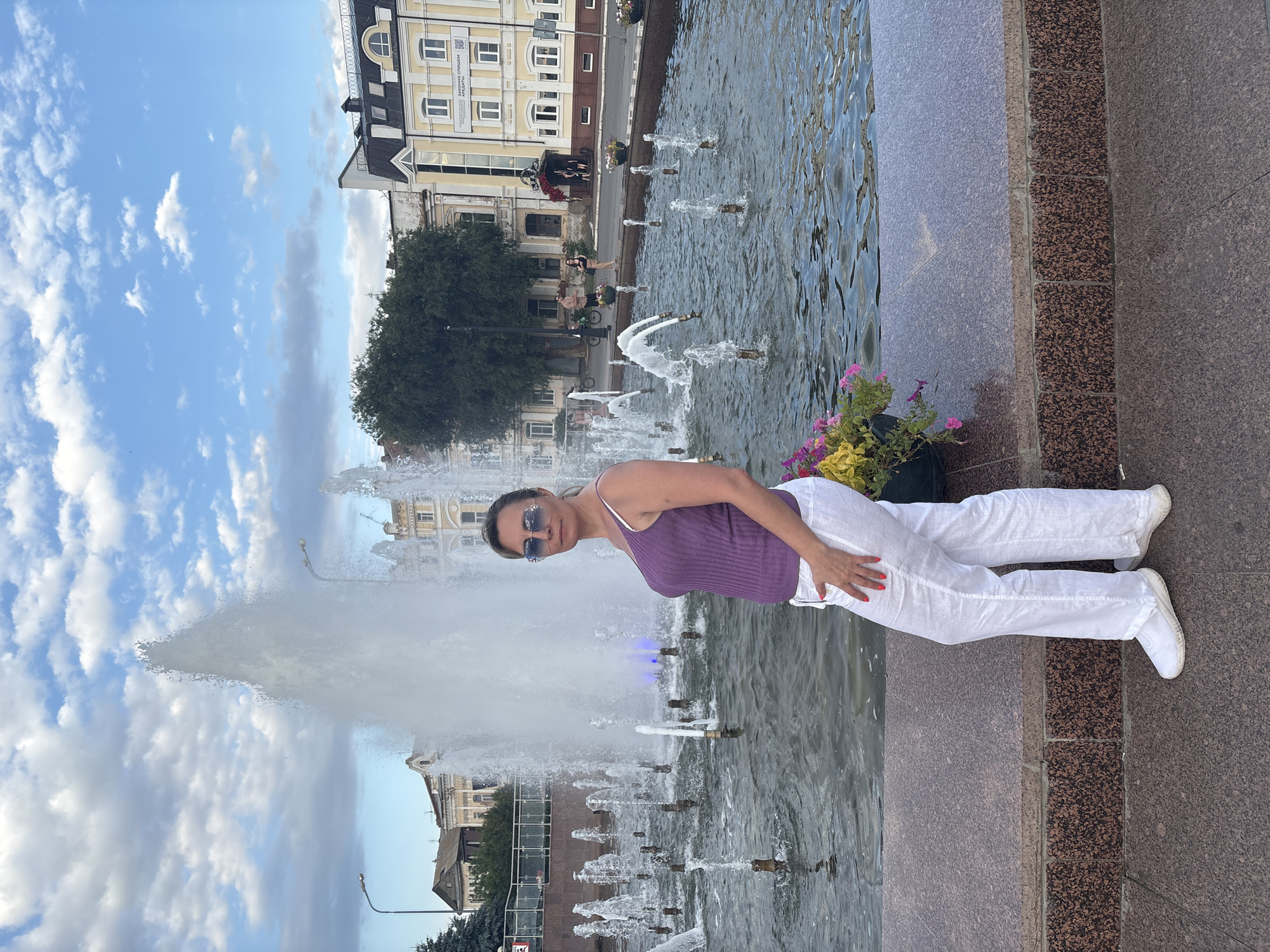 A woman stands in front of a fountain in Astrakhan, Russia, located on the embankment of the Volga River. The fountain has multiple jets of water spraying upwards. The woman is wearing a purple tank top and white linen pants, with sunglasses on. She is posing for the camera. In the background, there are buildings, trees, and a cloudy sky. The fountain area is lined with granite. Flowers are planted in pots along the edge of the fountain. There are other people and bicycles in the distance.