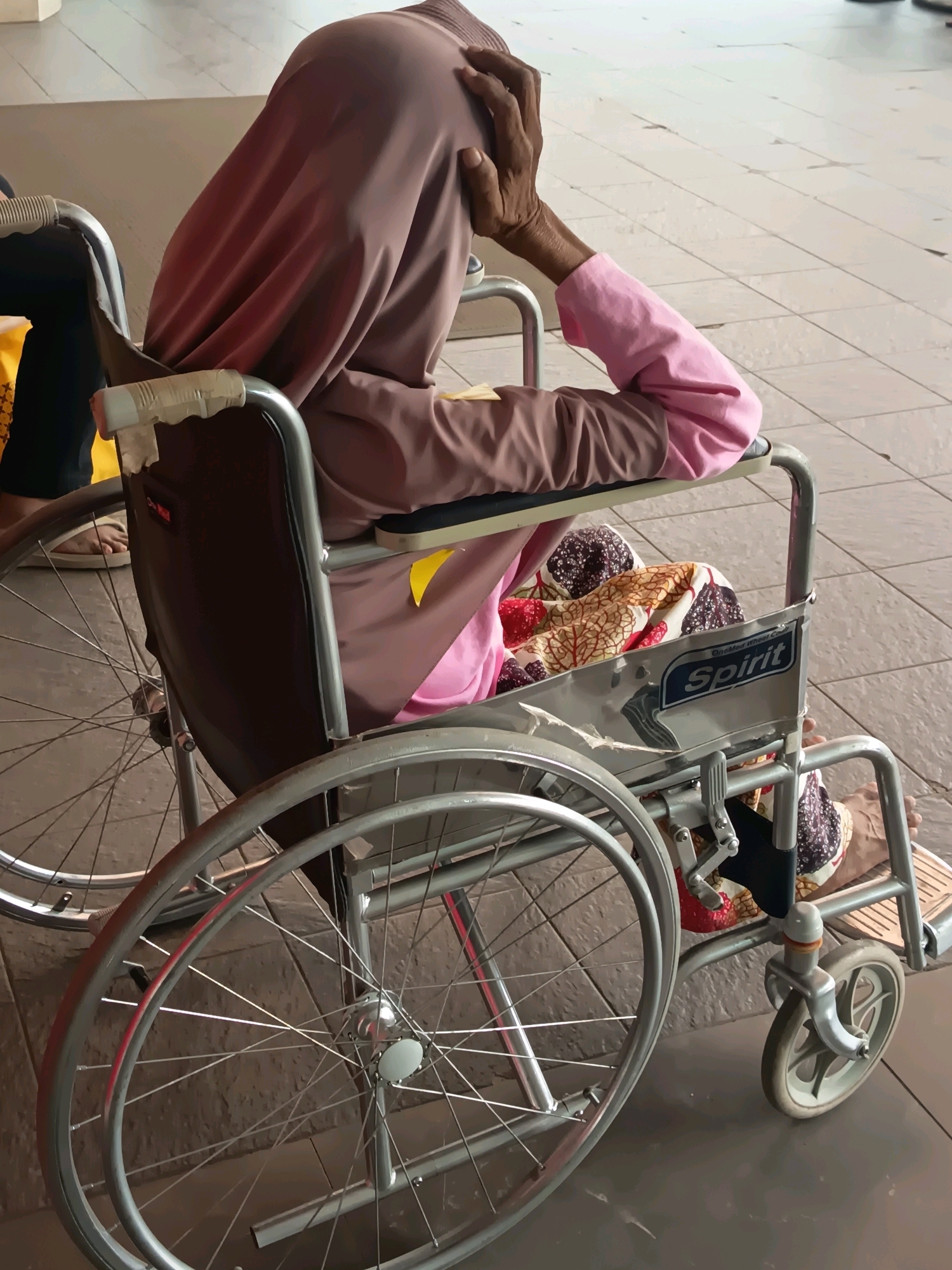 An elderly person wearing a pink hijab and a pink long-sleeved shirt is seated in a wheelchair. Their right hand is resting on their forehead, and their left arm is bent at the elbow, resting on the armrest. The wheelchair has a brown backrest with the word "Spirit" visible on a metallic plate. The person is covered with a patterned blanket featuring images of red and brown leaves on a white background. The scene appears to be indoors, possibly in a hospital or public waiting area, with a tiled floor in the background. The location is identified as Tangerang, Indonesia.