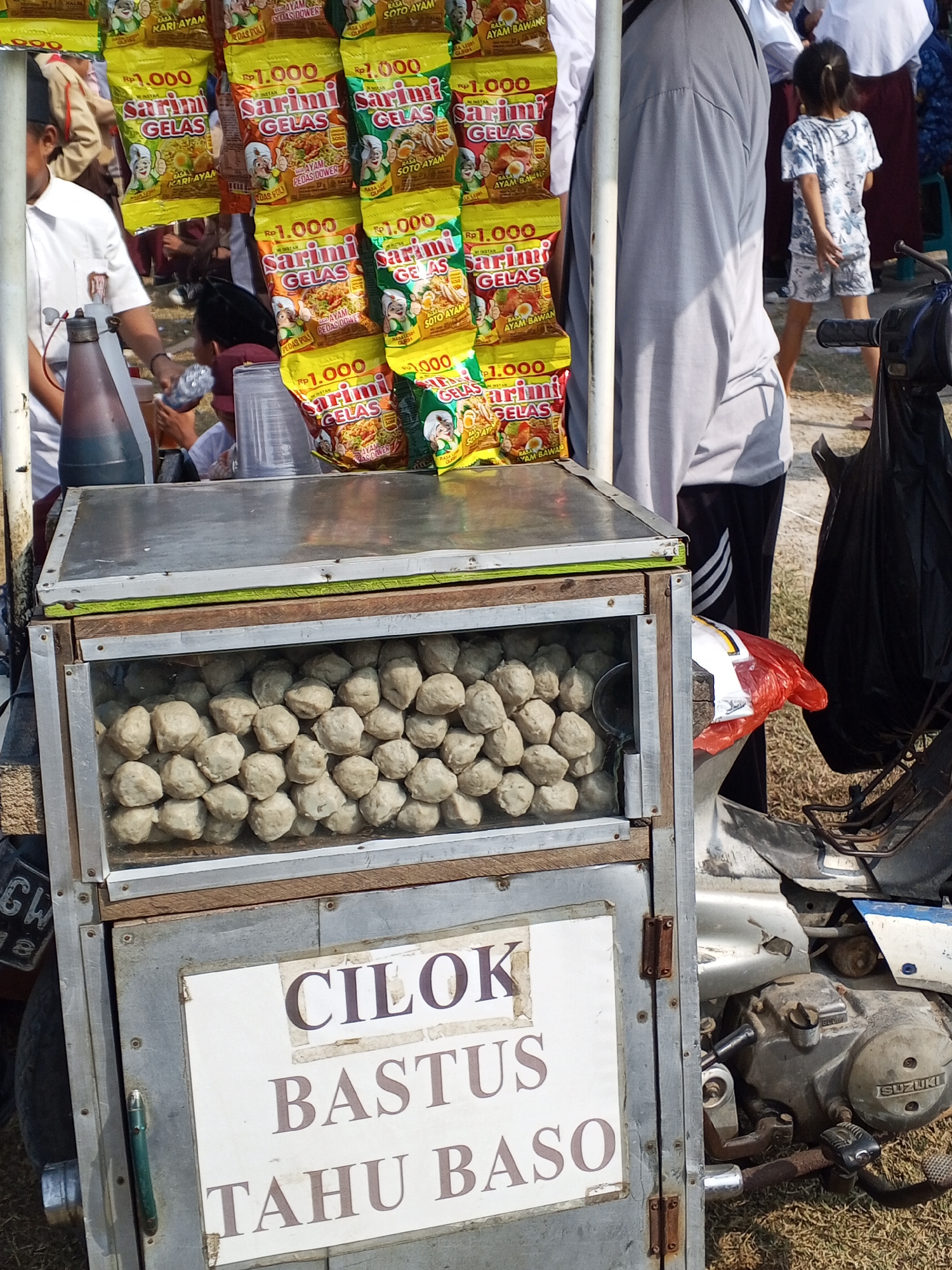 In Ranjeng, Indonesia, a street food cart displays an array of snacks. Packets of "Sarimi Gelas" instant noodles, priced at Rp 1.000, are prominently featured, with various flavors like "Rasa Ayam Bawang" and "Rasa Soto Ayam." Below the noodles, a glass-fronted compartment holds a large quantity of spherical meatballs, presumably "cilok" and "tahu baso," as indicated by text on the cart. The cart is attached to a motorcycle, suggesting mobility. In the background, several individuals, including a young girl in a patterned outfit and a person in a grey t-shirt, are visible. The setting appears to be outdoors, with dry grass on the ground.