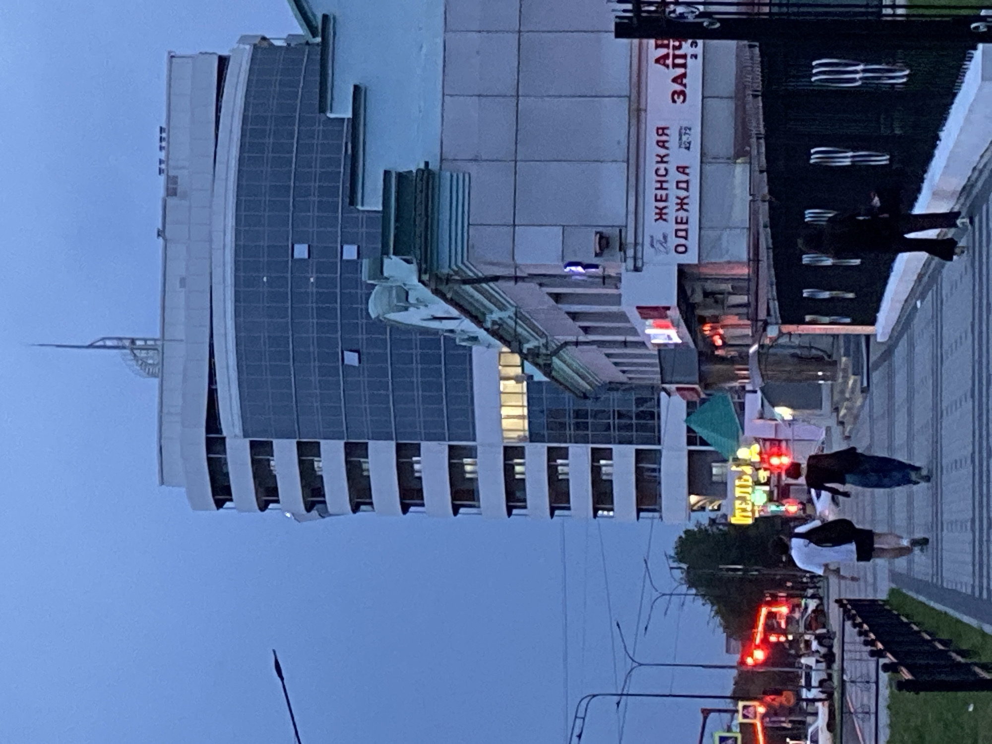 An urban street scene unfolds at dusk in Blagoveshchensk, Russia. A distinctive modern, curved building with multiple balconies and a glass facade dominates the background, contrasting with older, lower-rise structures in the foreground. A pedestrian walkway, paved with rectangular stones, leads towards the viewer, bordered on the right by a black metal fence. Two individuals, viewed from behind, walk away from the camera on the walkway. The person on the left wears a white t-shirt and black shorts, carrying a black bag over their shoulder. The person on the right is dressed in dark clothing and dark flared pants. The street to the left features traffic lights, illuminated signs, and parked vehicles. Signs on the building in the foreground include "ЖЕНСКАЯ ОДЕЖДА" (Women's Clothing) and "АПТЕКА" (Pharmacy). The overall lighting suggests late evening or early morning.
