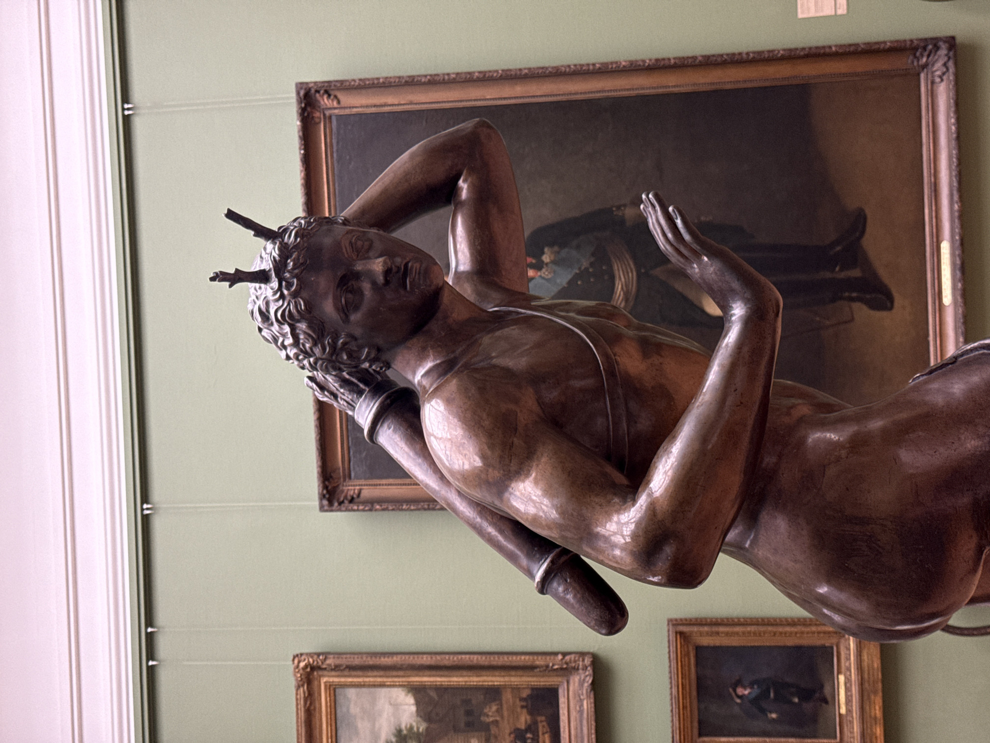 The image depicts a bronze sculpture of a reclining male figure with stag antlers protruding from his head. He has a muscular build, curly hair, and appears to be resting on his arm. His other hand is raised, fingers slightly spread. The sculpture is on display in a museum or gallery setting in Moscow, Russia. Behind the sculpture are three framed paintings hanging on a pale green wall, with a white door frame visible on the left. Hanging wires are seen on the wall, likely used for the framed art.