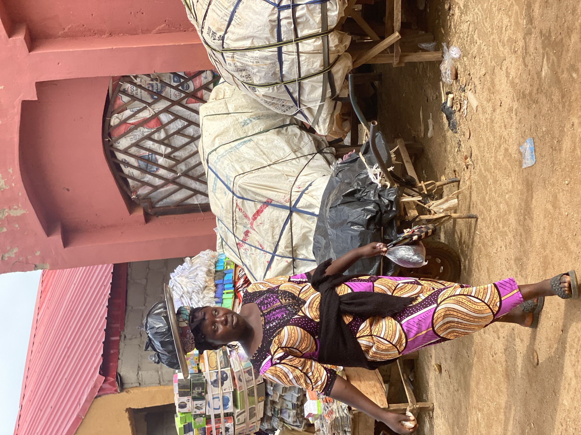 A woman stands in a street market in Kafanchan B, Nigeria. She is wearing a purple and orange patterned dress with a black sash and sandals. She is holding food in plastic bags in her hands and has a tray balanced on her head loaded with goods. In the background, there are buildings, sacks of goods on a cart, and other market stalls. The ground is dirt.