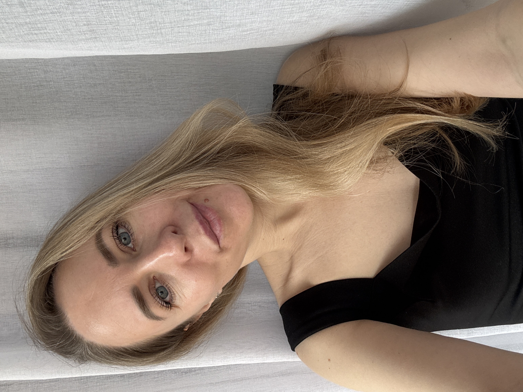 A woman with blonde hair and blue eyes is lying down, facing the camera. She is wearing a black off-the-shoulder top. The background is a textured white fabric, likely a sheet or curtain. No evidence indicates this photo was taken in Chaikovsky, Russia.