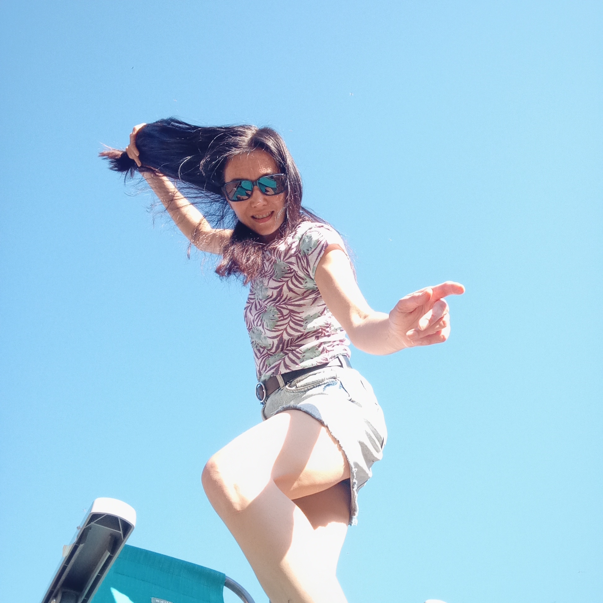 A low-angle shot shows a woman with dark hair and sunglasses standing on a turquoise beach chair, with a clear blue sky as the background. She is wearing a light-colored short-sleeved shirt with a leaf pattern and denim shorts. Her hair is partially pulled back, and she is gesturing with her right hand pointed forward. The location is not discernible.