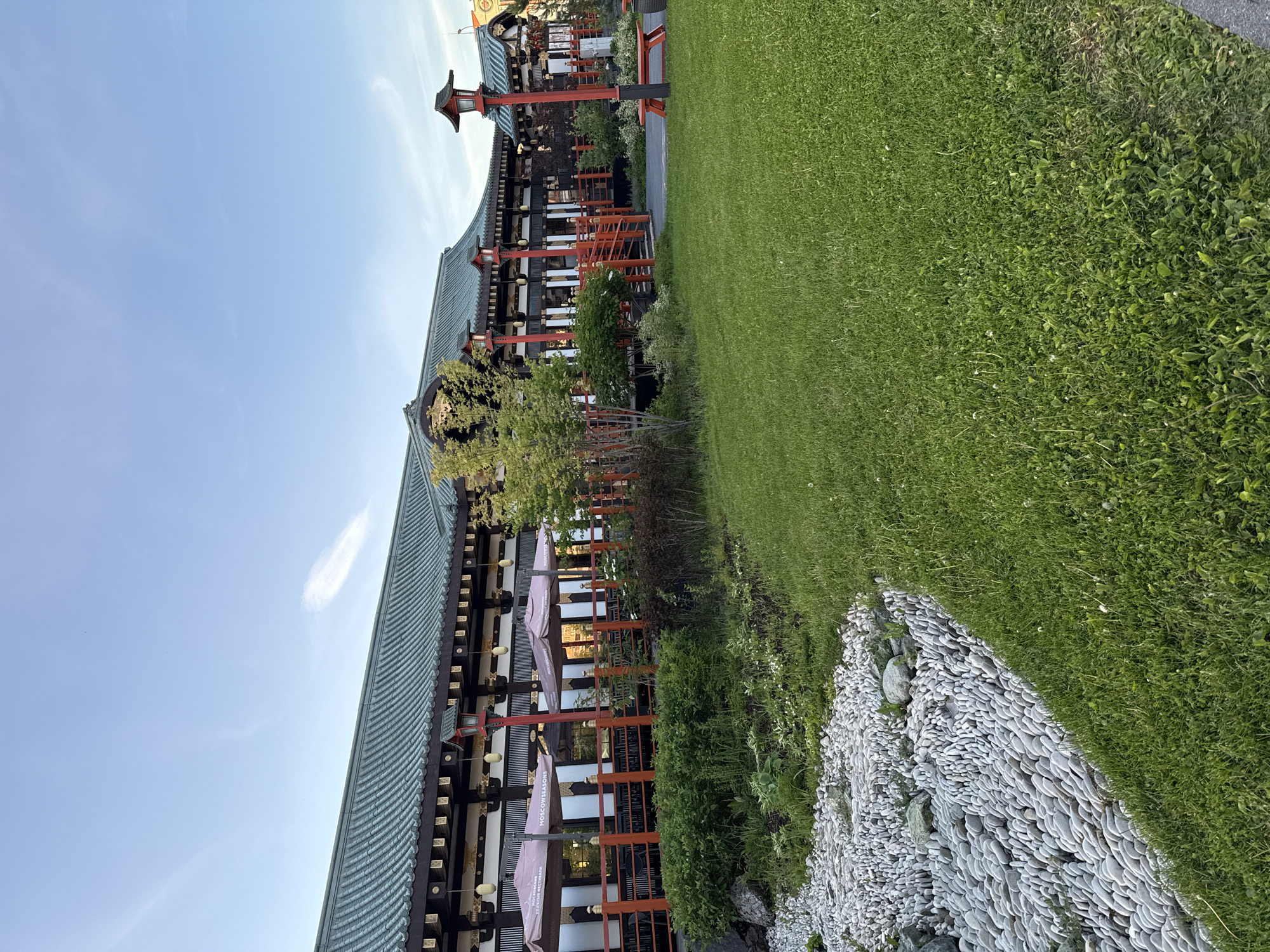 This image captures a serene outdoor scene featuring a building with distinct East Asian architecture, characterized by its multi-tiered, dark-tiled roof, ornate eaves, and prominent red structural elements. The building, possibly a restaurant, has an outdoor dining area equipped with several tables and light-colored patio umbrellas. Red lanterns or lights hang from the eaves and are mounted on red posts, enhancing the traditional aesthetic.
In front of the building, a narrow garden bed filled with lush green shrubs and plants separates it from a neatly manicured green lawn. A unique decorative feature is a border between the lawn and the garden bed, made of numerous flat, light-colored stones arranged in an overlapping, scale-like pattern. The sky above is clear and light blue with a few faint clouds, indicating daytime, likely late afternoon due to the soft lighting.
No people are visible, lending a tranquil atmosphere to the scene. The overall impression is one of cultural elegance and peaceful outdoor dining. A notable detail is the text visible on one of the patio umbrellas, which clearly reads "MOSCOW SEASONS," and possibly "RESTAURANT" below it. This suggests the establishment is a themed restaurant located in Moscow, despite the architectural style strongly evoking East Asia.