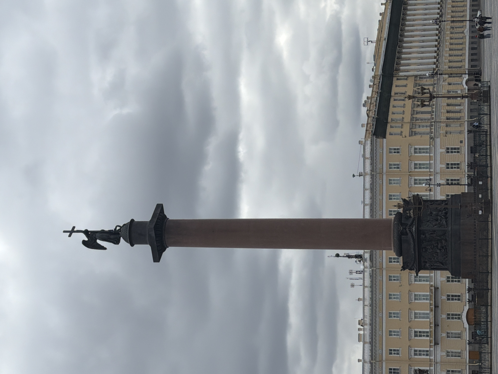 The image shows the Alexander Column in Palace Square, Saint Petersburg, Russia. The column is a tall, reddish-brown monolithic granite structure topped with a bronze statue of an angel holding a cross. The base of the column features intricate bronze reliefs. Behind the column is the Winter Palace, a large yellow building with rows of windows and white columns. The sky is overcast with gray clouds. There are several people walking in front of the Winter Palace. The scene appears to be taken from ground level, looking up at the column and building.