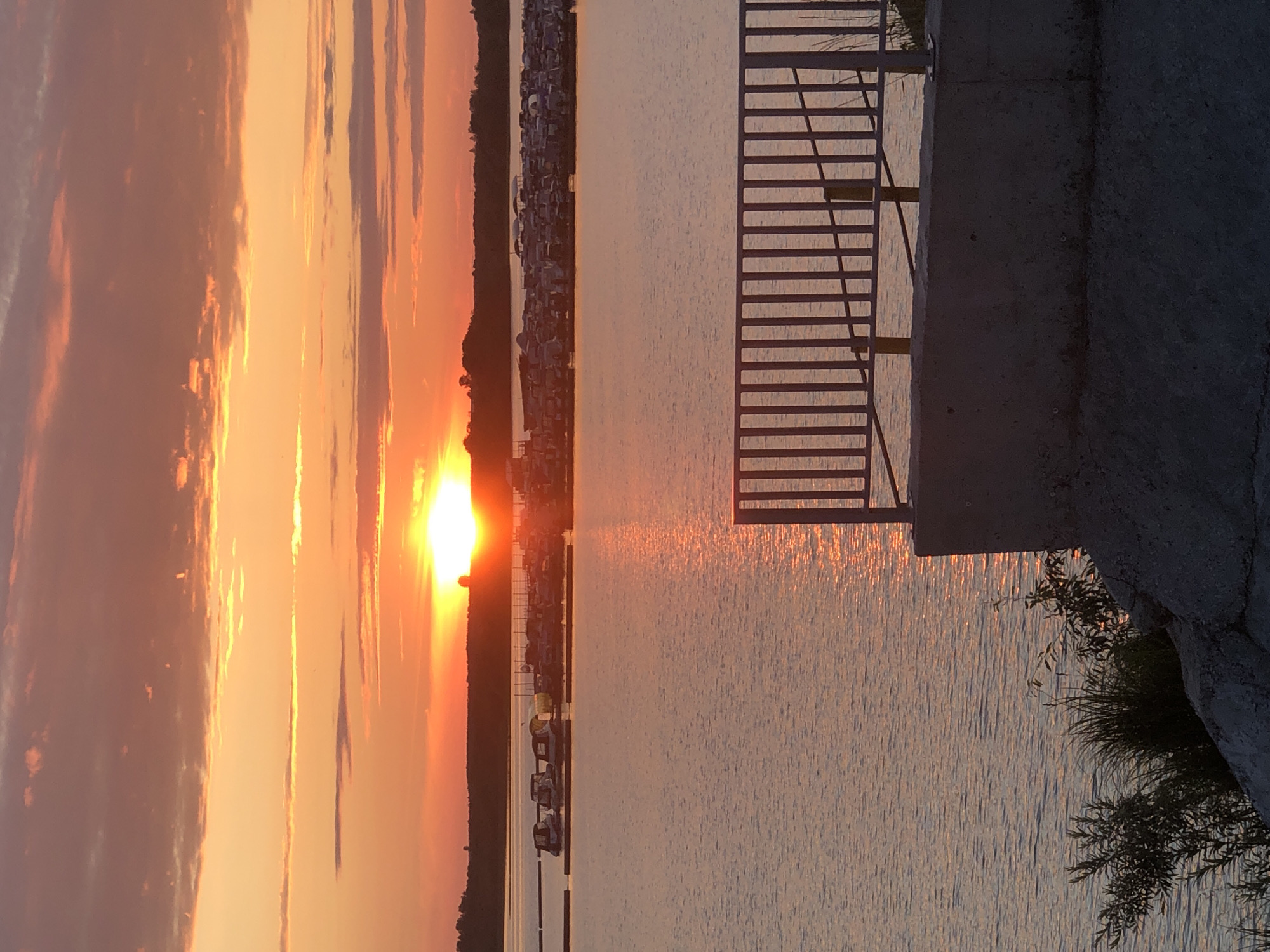 The image depicts a sunset over a body of water, possibly a river or lake, with a coastline lined with trees and structures. The sun is low on the horizon, casting a warm, orange glow across the sky and reflecting on the water's surface. Clouds are present, tinged with the colors of sunset. On the right side of the image, a concrete structure with a metal railing is visible, suggesting a pier or waterfront area. The railing casts a shadow on the water. The scene conveys a sense of tranquility and natural beauty, characteristic of a sunset. The lighting and colors suggest it is either early morning or late evening. The location context provided is Solikamsk, Russia, which helps to identify the setting as a specific geographical area. No people or text are visible in the image.