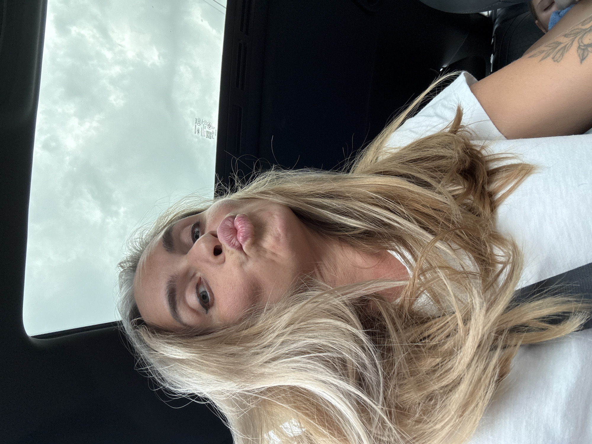 A blonde woman with her hair loose is leaning forward into the frame, making a kissy face. She is wearing a white t-shirt and a grey seatbelt is visible across her chest. The background is dark, suggesting the interior of a vehicle, with a sunroof showing a cloudy sky. A tattoo is visible on her right arm. The image is a close-up selfie.