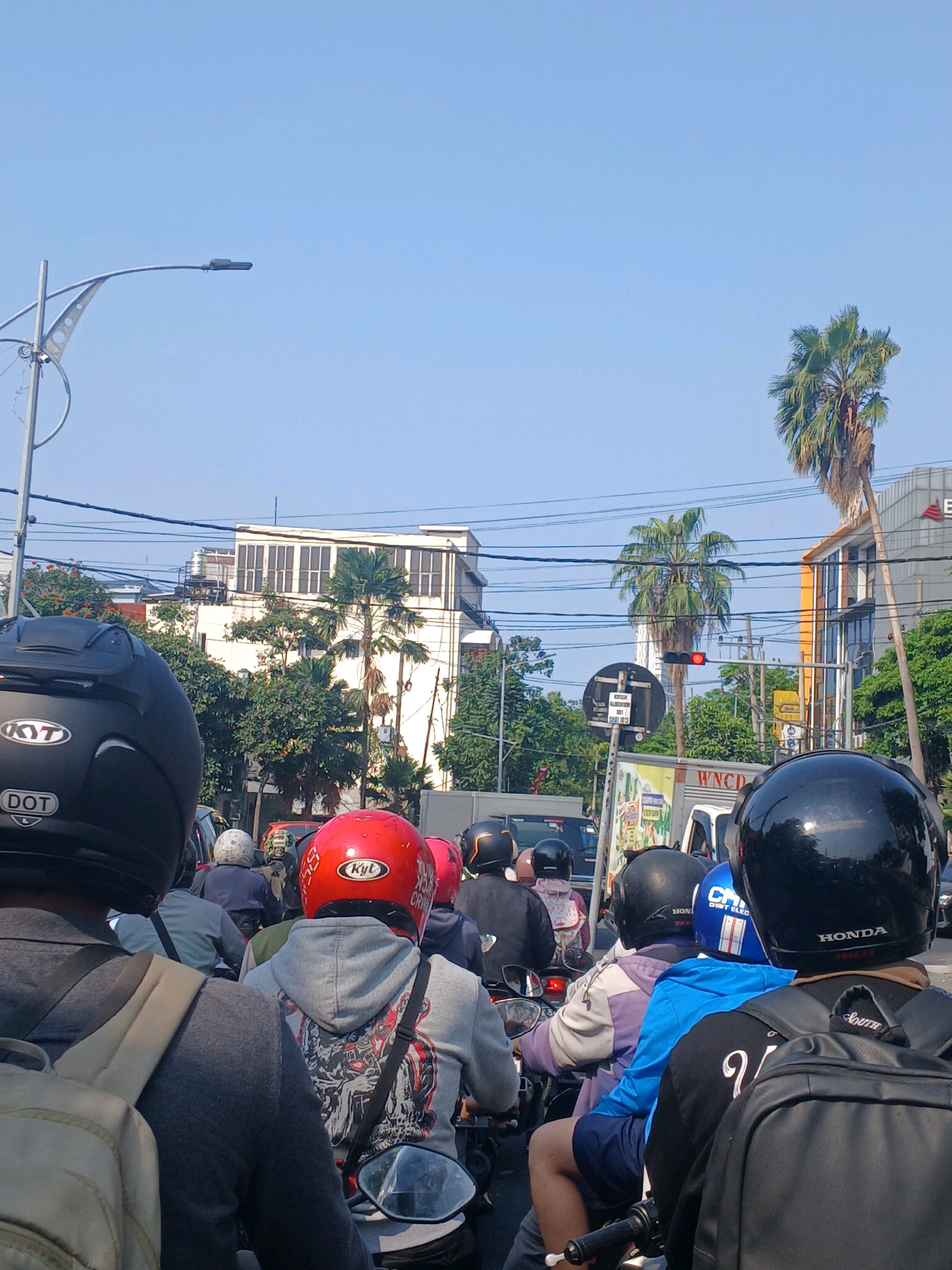 The image depicts a traffic scene in Surabaya, Indonesia, likely on a street with moderate to heavy vehicle congestion. The perspective is from the back of a motorcycle, looking forward at a line of other motorcycles and their riders. The sky is clear and blue, suggesting a daytime scene with sunny weather.
In the foreground, the backs of several individuals wearing helmets are visible. The rider in the leftmost position wears a black helmet with "KYT" and "DOT" markings. Next to them is another rider with a gray hoodie and a distinctive graphic design on the back. Further ahead, a rider in a bright red helmet with "KYT" branding is prominent. Other riders are seen with helmets of various colors, including black and blue. Many riders are wearing casual clothing such as t-shirts and hoodies, and some have backpacks.
The environment includes elements of an urban landscape. Tall palm trees are visible, a common sight in tropical cities like Surabaya. Buildings of varying architectural styles are present, including a white building with a grid-like facade and a more modern building with orange accents. Power lines crisscross the sky, indicating active infrastructure. A traffic light displaying a red signal is visible, contributing to the traffic jam. A white truck with "WNCD" printed on its side is also part of the traffic flow. The overall impression is a snapshot of daily life and transportation in a busy Indonesian city.