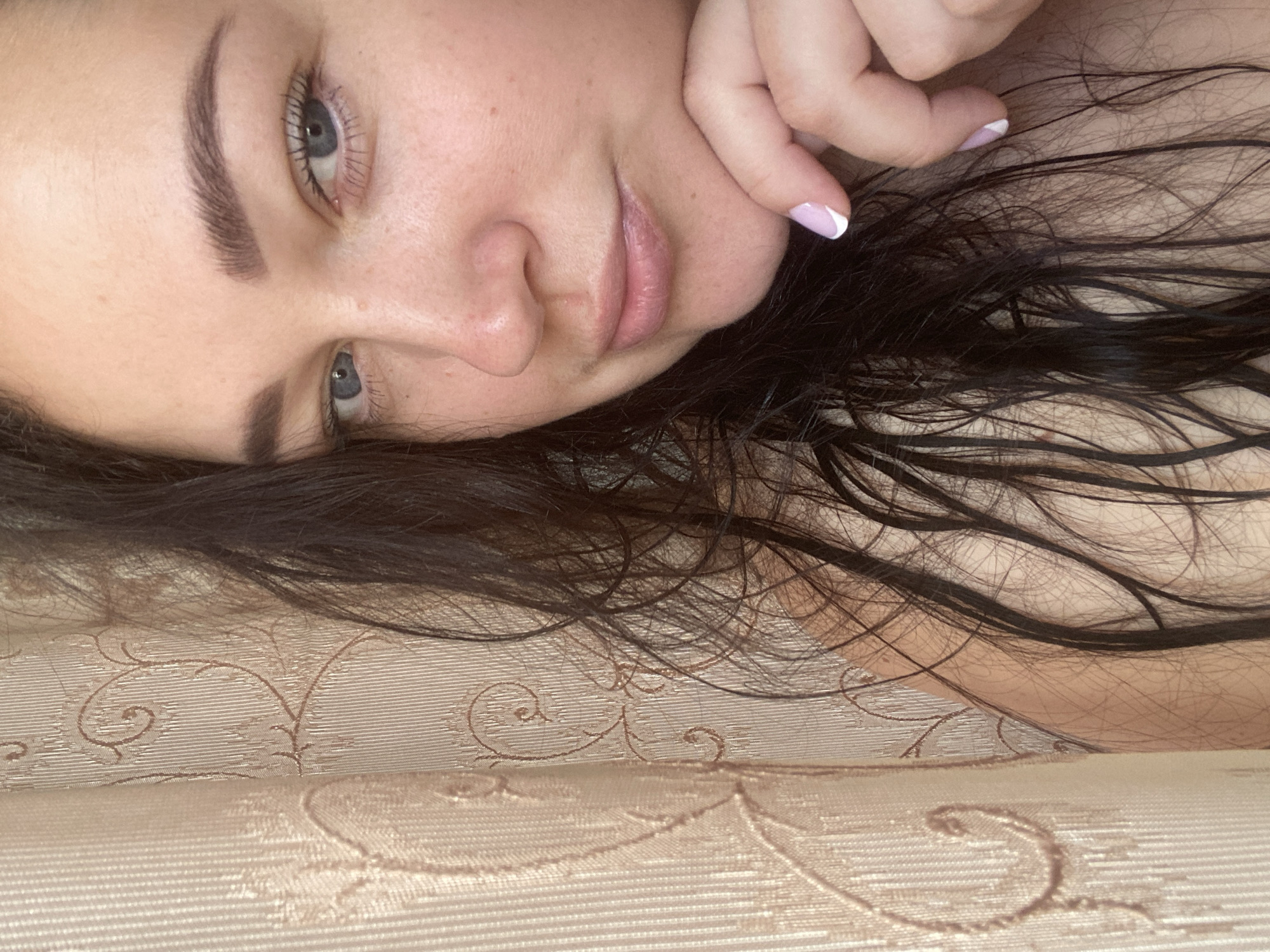 A close-up, medium-shot portrait of a woman with fair skin, light blue eyes, and dark brown hair. She is lying on a light-colored, textured surface, possibly fabric or upholstery with a subtle pattern. Her hair is spread across the surface. She has manicured nails with a white tip design on her left hand. The setting cannot be determined based on the image. The location in the city of Sochi, Russia is unknown.