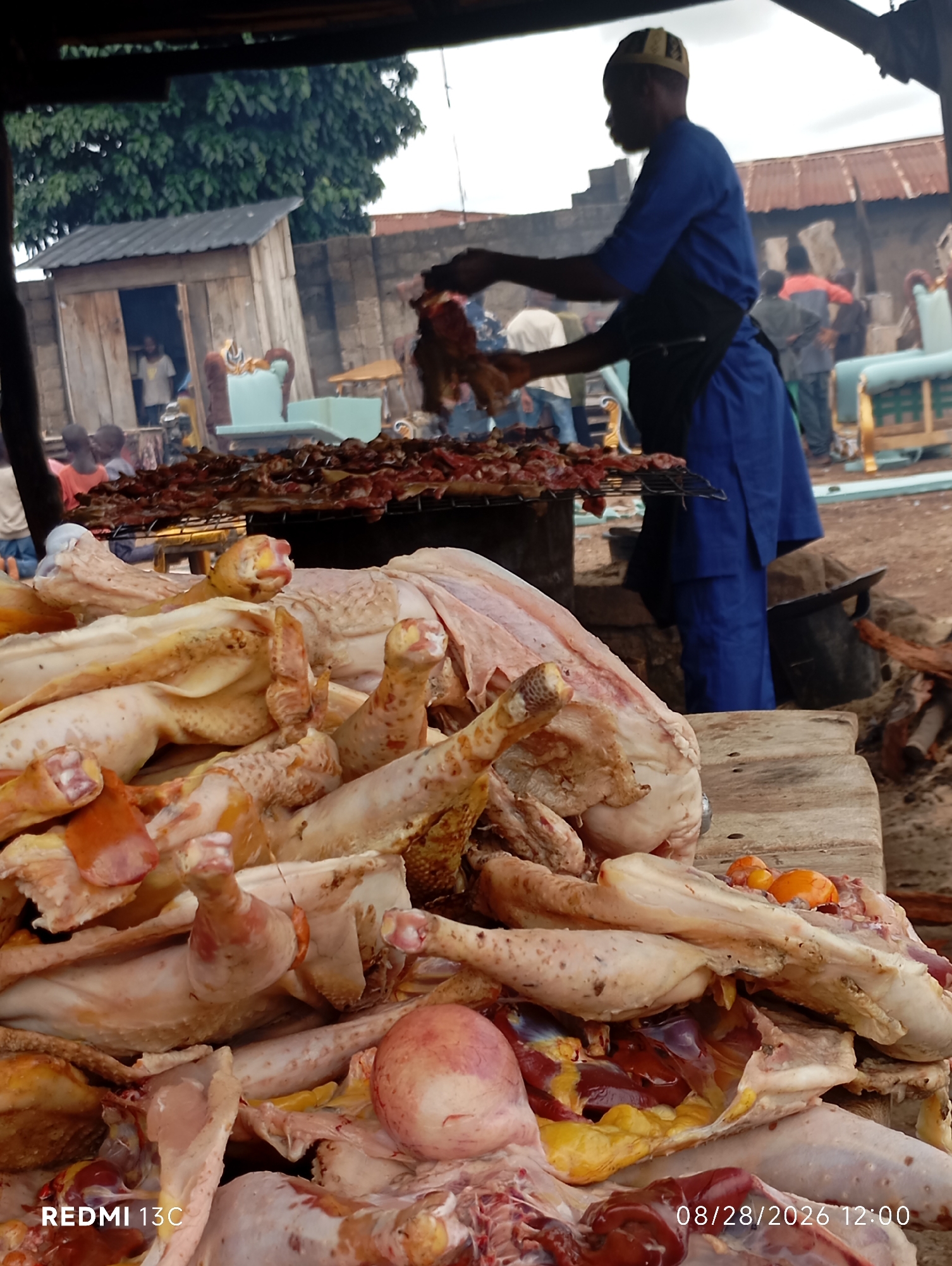 In Shendam, Nigeria, a man in a blue outfit and a black apron is seen at an outdoor food stall. He is actively preparing raw chicken, with a pile of prepared chickens in the foreground. The chickens are mostly whole, some with visible internal organs and eggs. Behind the man, a grill with meat cooking is visible, suggesting a cooking or preparation area. Other individuals are present in the background, some appearing to be customers or workers. The environment seems to be a busy marketplace or street food vendor setting, with simple wooden structures and roofing visible. The overall scene captures a moment of food preparation and commerce in a local Nigerian community.