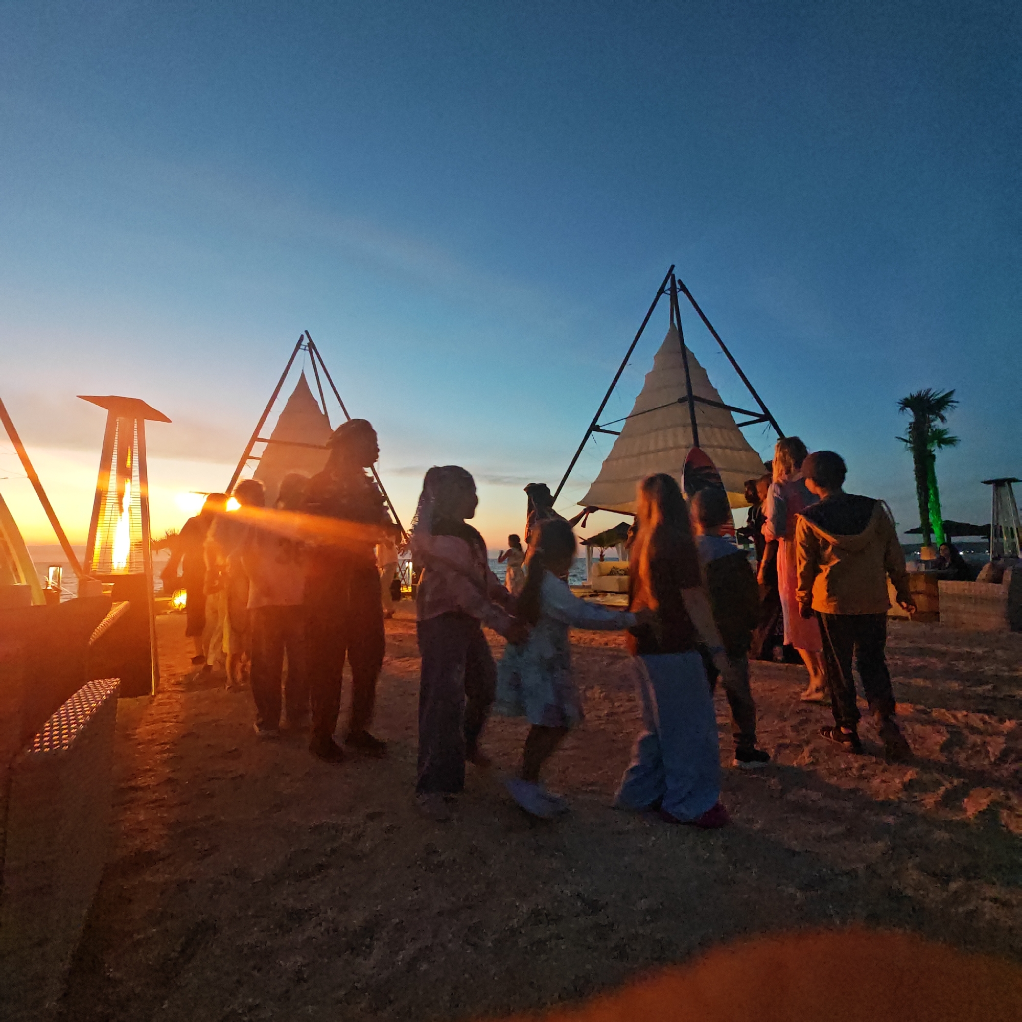 The image depicts an outdoor gathering at dusk, possibly at a beach or waterfront, given the presence of sand and the ocean in the background. The sky is a gradient of blue and orange, indicating sunset. Several people, including adults and children, are visible, some of whom appear to be dancing or interacting in a group. Two conical tent-like structures with white fabric are prominent in the background, suggesting a festive or event setting. To the left, a tall outdoor heater with a visible flame adds to the ambient lighting. A palm tree is visible on the right side of the image. The overall atmosphere appears relaxed and celebratory. The location is identified as Мысовское сельское поселение, Ukraine.