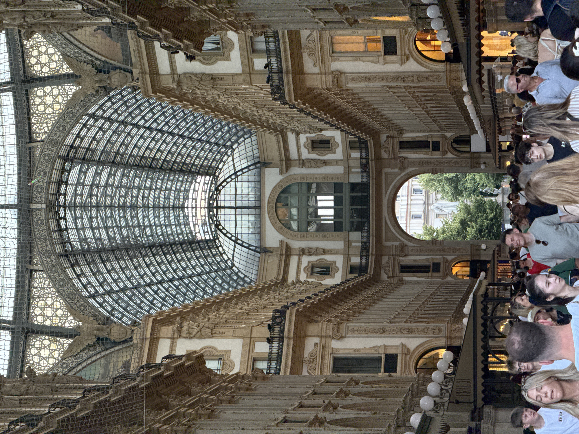 The image shows the Galleria Vittorio Emanuele II in Milan, Italy, a famous 19th-century glass-vaulted shopping arcade. The perspective is from within the arcade, looking down its length towards an archway opening to an outdoor area. The arcade's architecture is characterized by ornate facades, arched windows, and intricate detailing, all topped by a large, curved glass roof supported by a metal framework. Sunlight streams through the glass roof, illuminating the interior. A crowd of people is visible, some walking and others standing, suggesting a bustling environment. Shops and cafes are situated along the sides of the arcade, with awnings and glowing lights indicating their presence. The overall scene depicts a vibrant and architecturally significant public space in Milan.