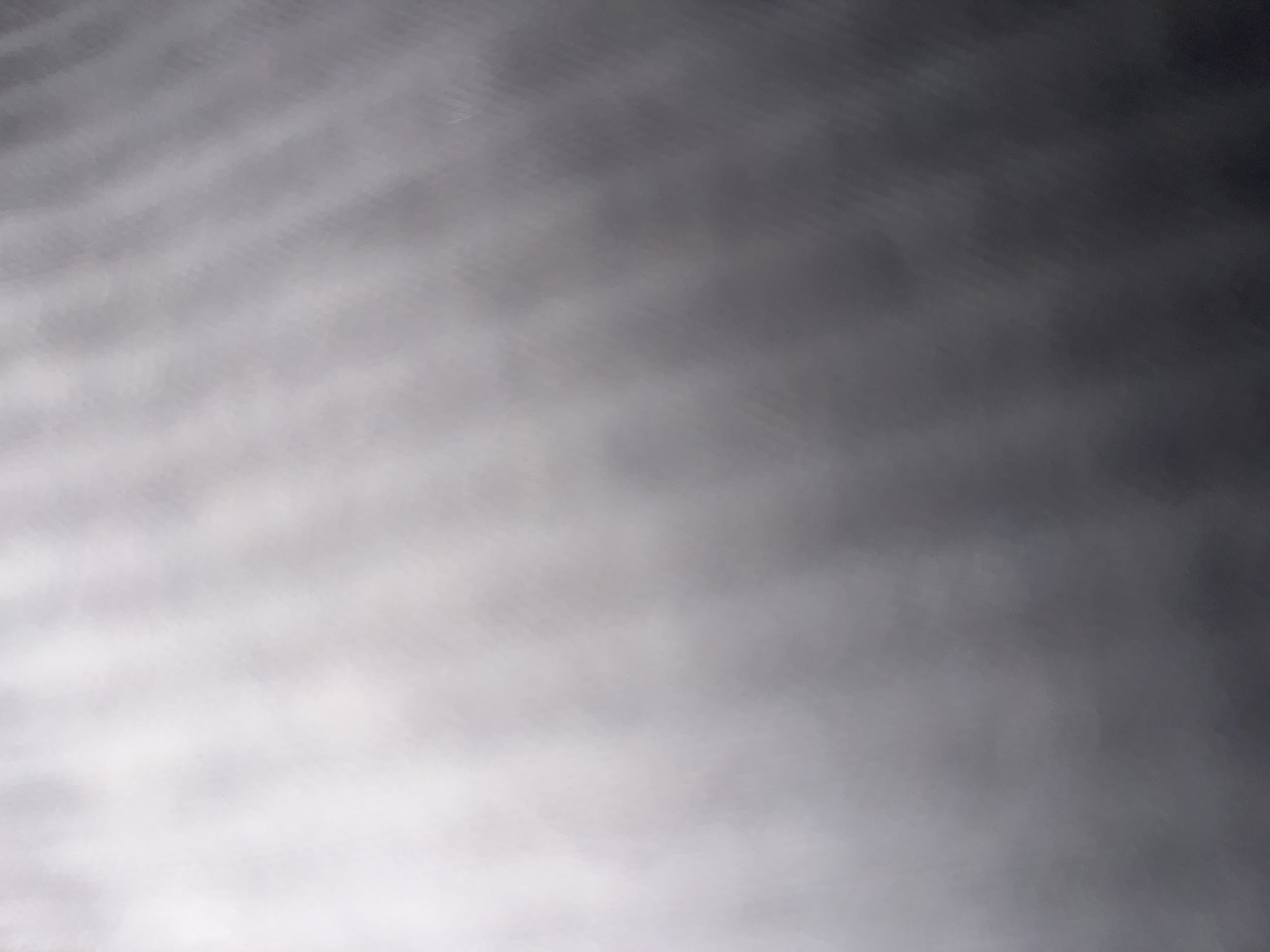 An abstract, out-of-focus image displays a gradient of light and shadow. The upper left portion is a bright, hazy white, gradually transitioning to a darker grey and black towards the lower right. A subtle, textured pattern of diagonal lines or waves is visible throughout the darker areas, suggesting a material or surface with some form of texture. There are no discernable objects, people, or specific environmental features. The image appears to be a close-up or macro shot of an abstract texture.