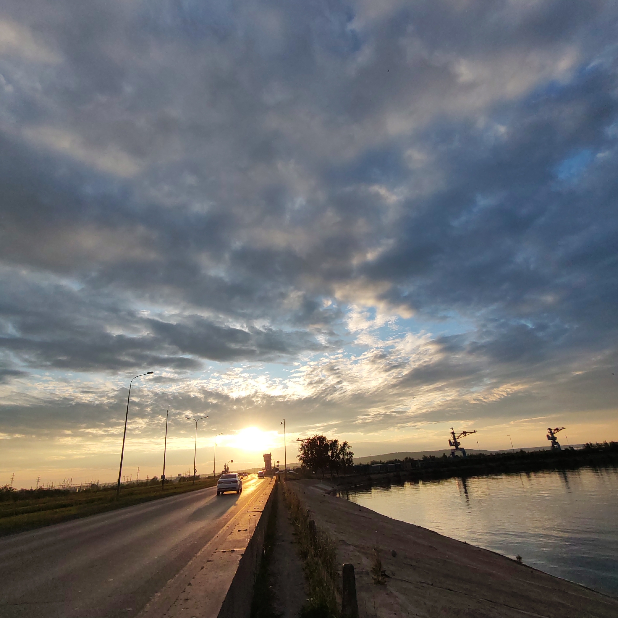The image depicts a road running alongside a body of water, likely the Kama River, on the outskirts of Chaikovsky, Russia, during sunset. The sky is filled with dramatic clouds, ranging from dark gray to lighter blues and whites, illuminated by the setting sun. The sun itself is a bright orb peeking through the clouds, casting a golden glow on the scene. On the left side of the image, a road is visible with a few cars driving on it. Streetlights line the side of the road. The right side of the image shows a concrete embankment sloping down to the water, with some vegetation growing along its edge. In the distance, across the water, industrial cranes are visible against the horizon, suggesting a port or industrial area. The overall impression is one of a serene, yet industrial, landscape at dusk.